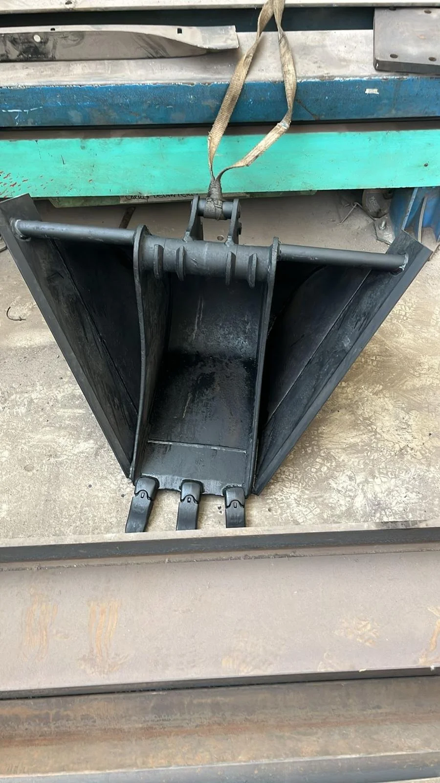 Cuchara excavadora de metal negra, vacía, con tres ganchos en la base, suspendida por una correa de tela en un taller.
