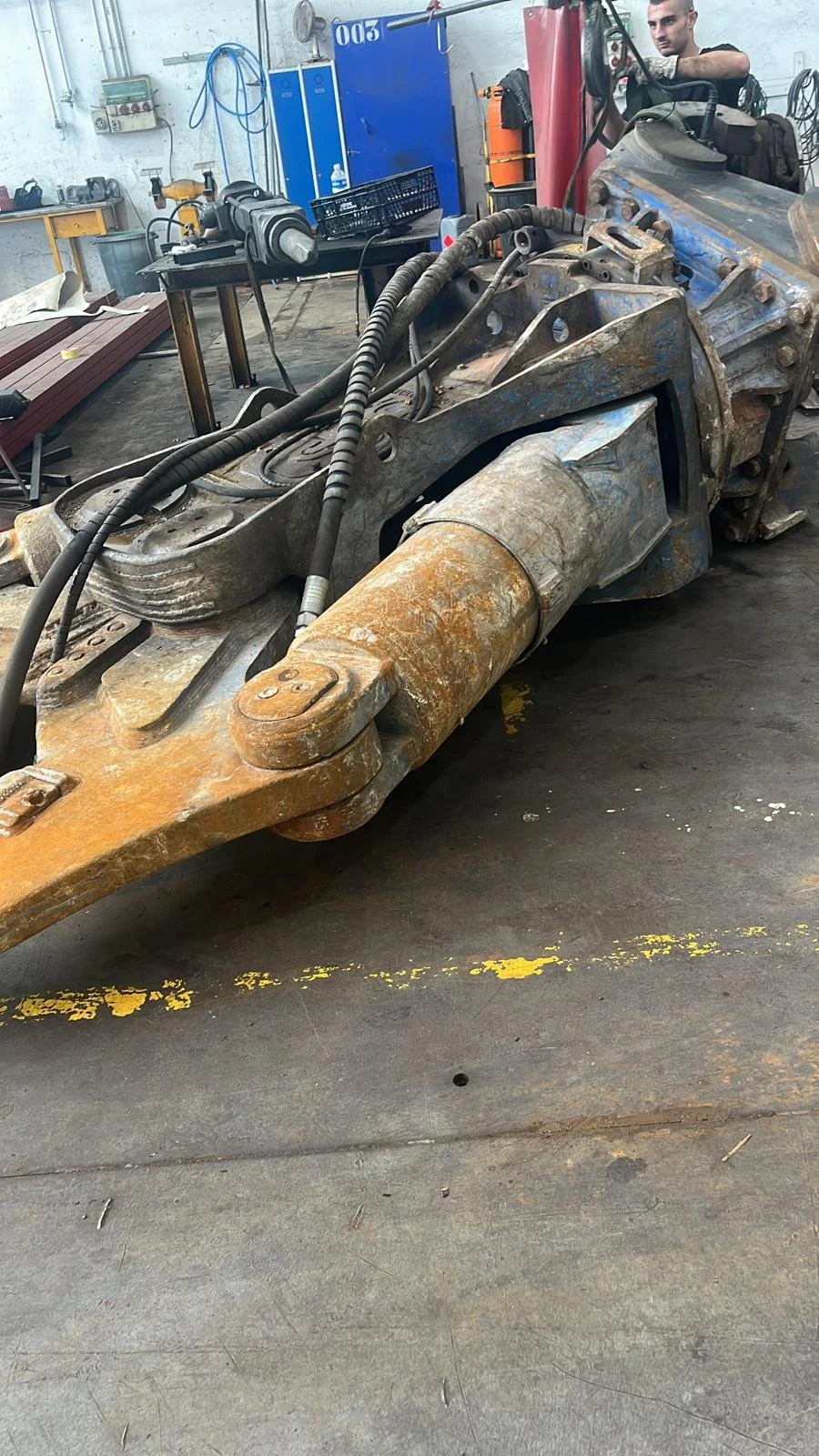 Una pieza de equipo de construcción grande y oxidada en un taller, con un hombre observando en el fondo, y herramientas y gabinetes azules en la pared atrás.