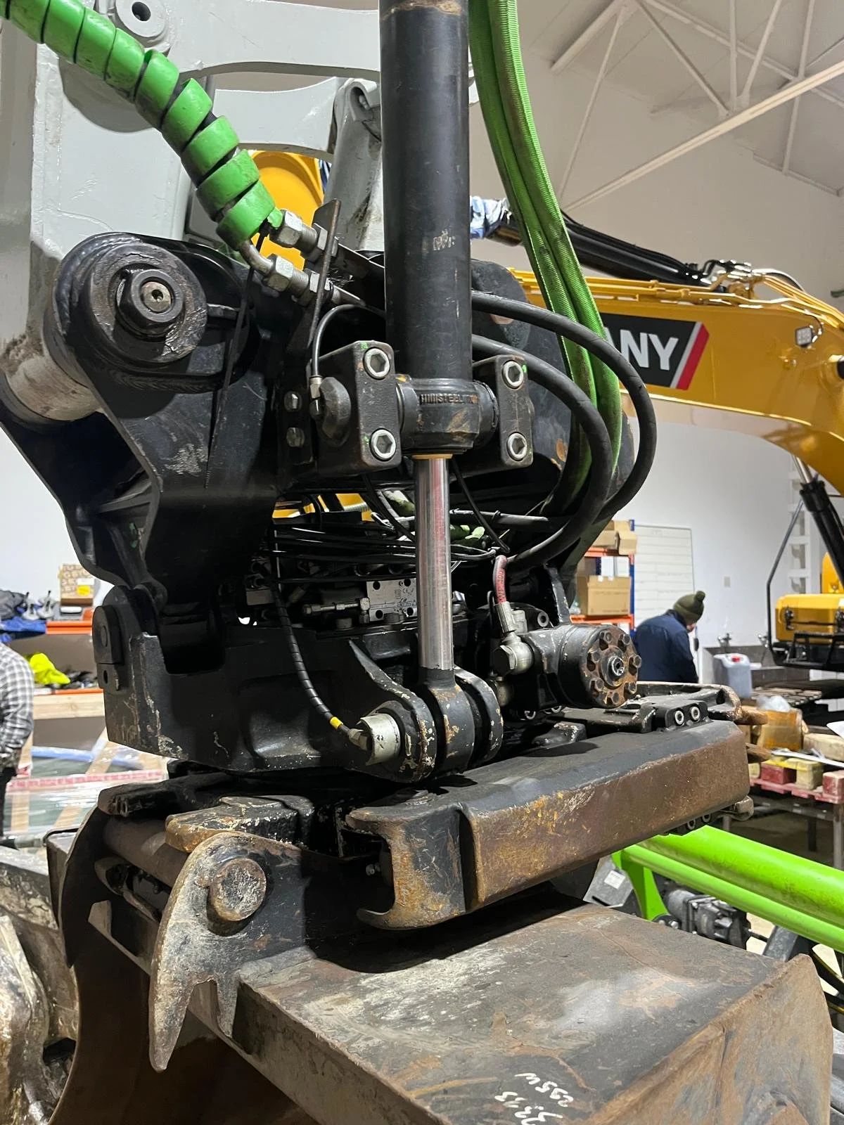 Componentes mecánicos de maquinaria pesada en un taller, con un vehículo de construcción y un brazo excavador visible en el fondo.