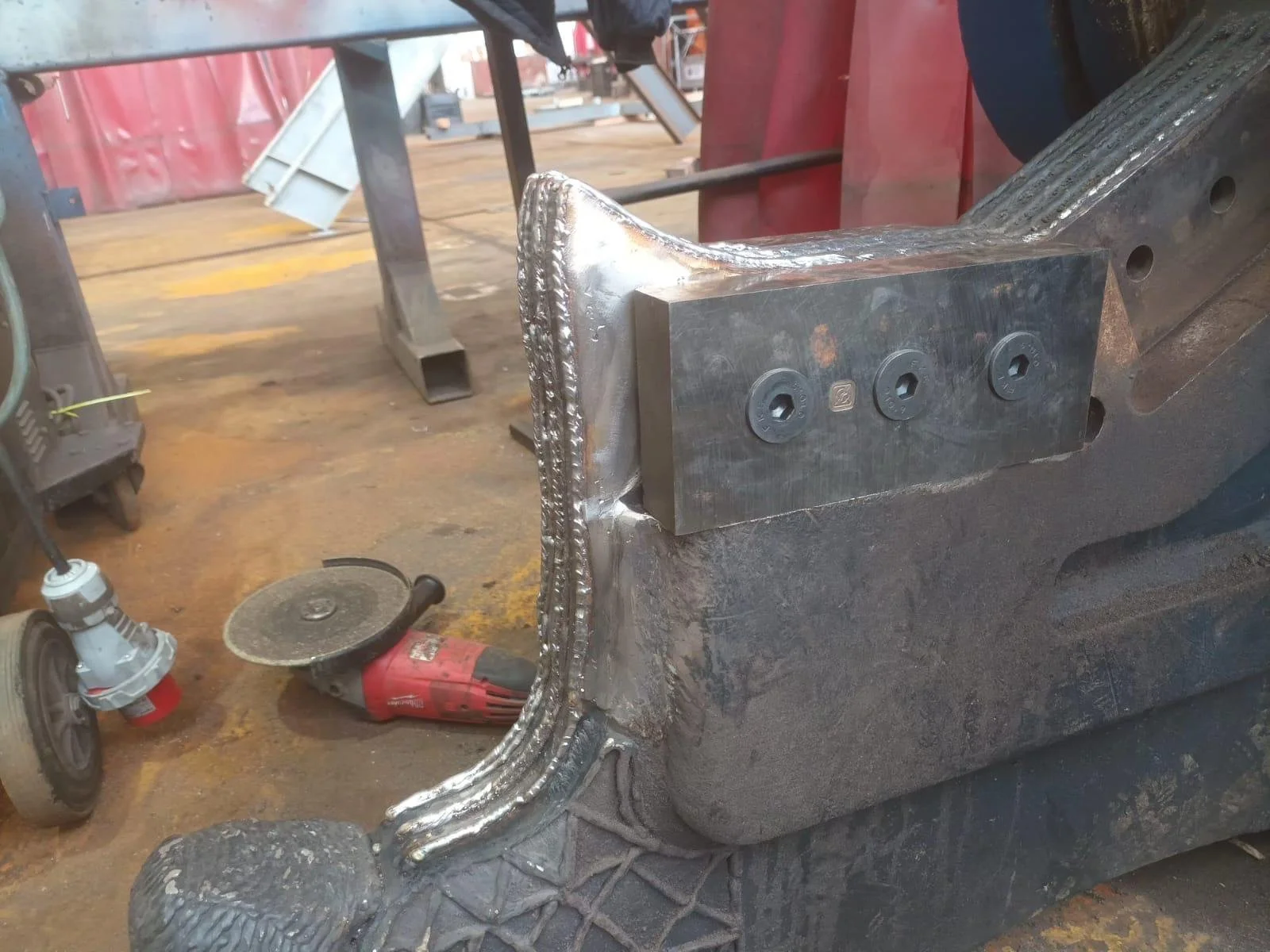 Una estructura de metal en proceso de soldadura en un taller. Se observa una pieza de metal siendo ensamblada con varias herramientas y partes de maquinaria a su alrededor.