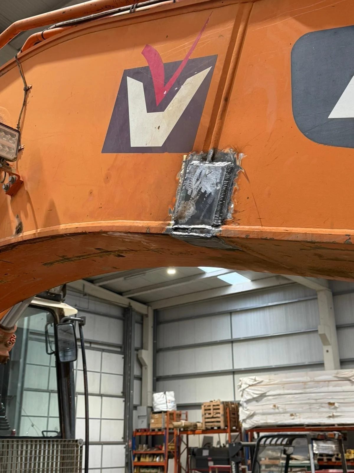 Parte superior de una máquina agrícola con un signo de verificación y una reparación de soldadura en el panel. En el fondo, un taller con estanterías y pallets de materiales.
