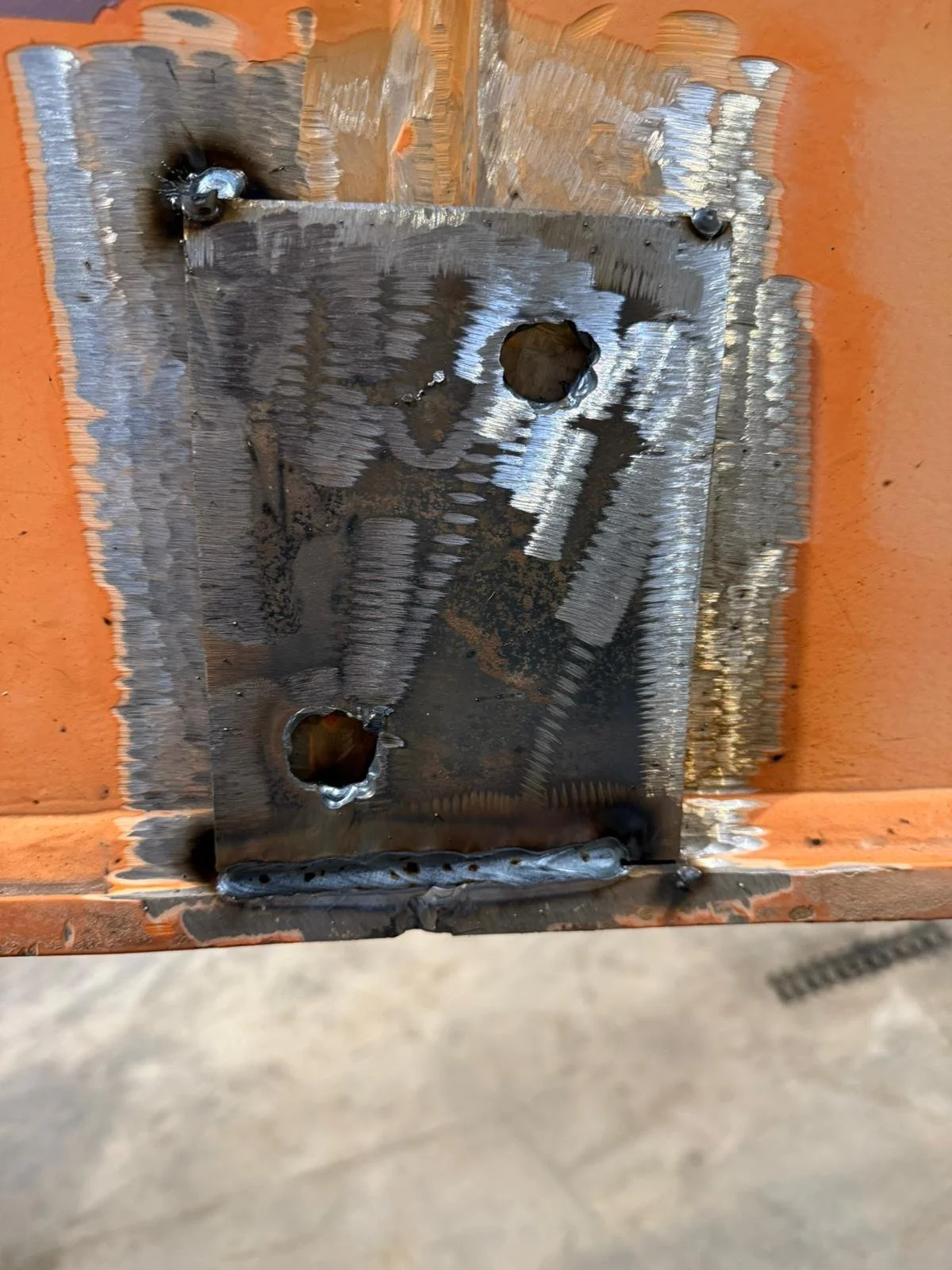 Placa de metal soldada a una superficie de madera, con dos agujeros rectangulares y una línea de soldadura en la parte inferior.