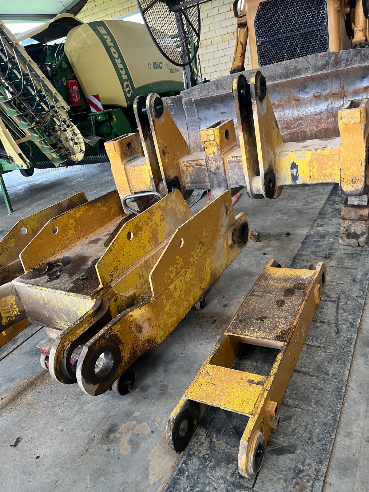 Partes de maquinaria agrícola amarillas y metálicas en un taller, con fondo de maquinaria grande y paredes de ladrillos.