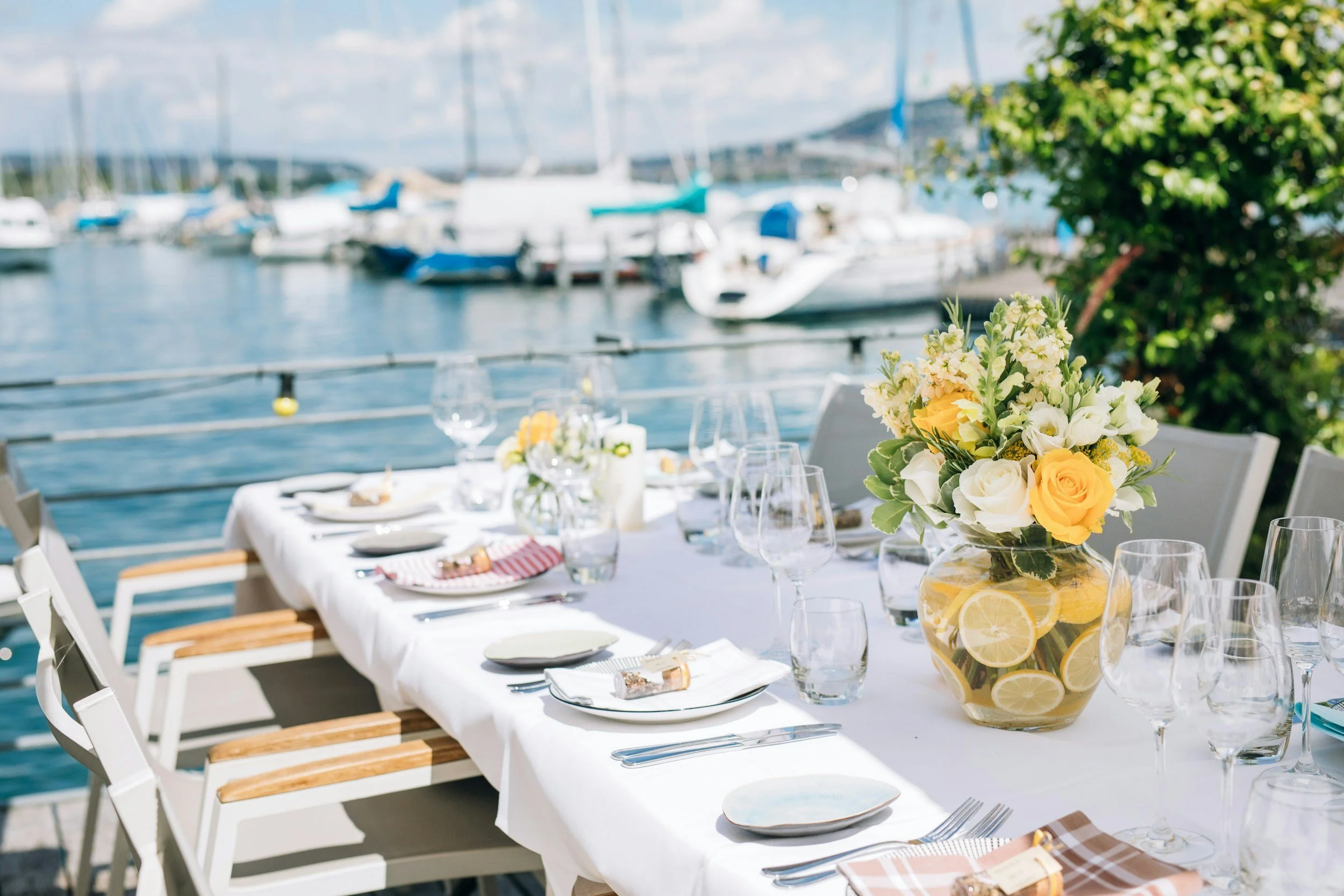 Dining table set outdoors near a marina with sailboats in the background, decorated with a floral arrangement in a lemon slice-themed vase, glassware, plates, and utensils.