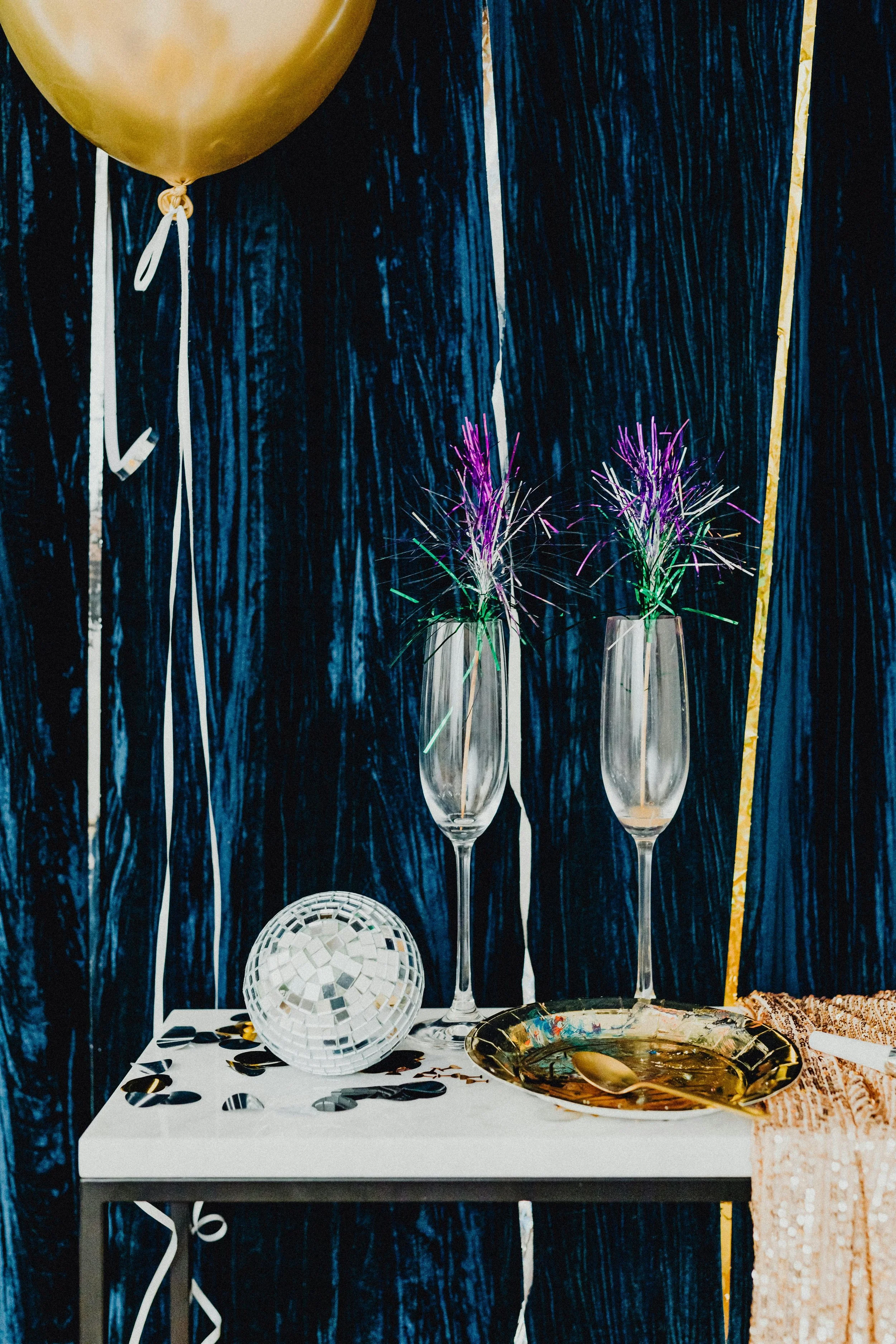 Decorative table with two champagne flutes holding colorful sparklers, a disco ball, a gold plate with a gold spoon, and party streamers, against a dark textured background with a gold balloon on the left side.