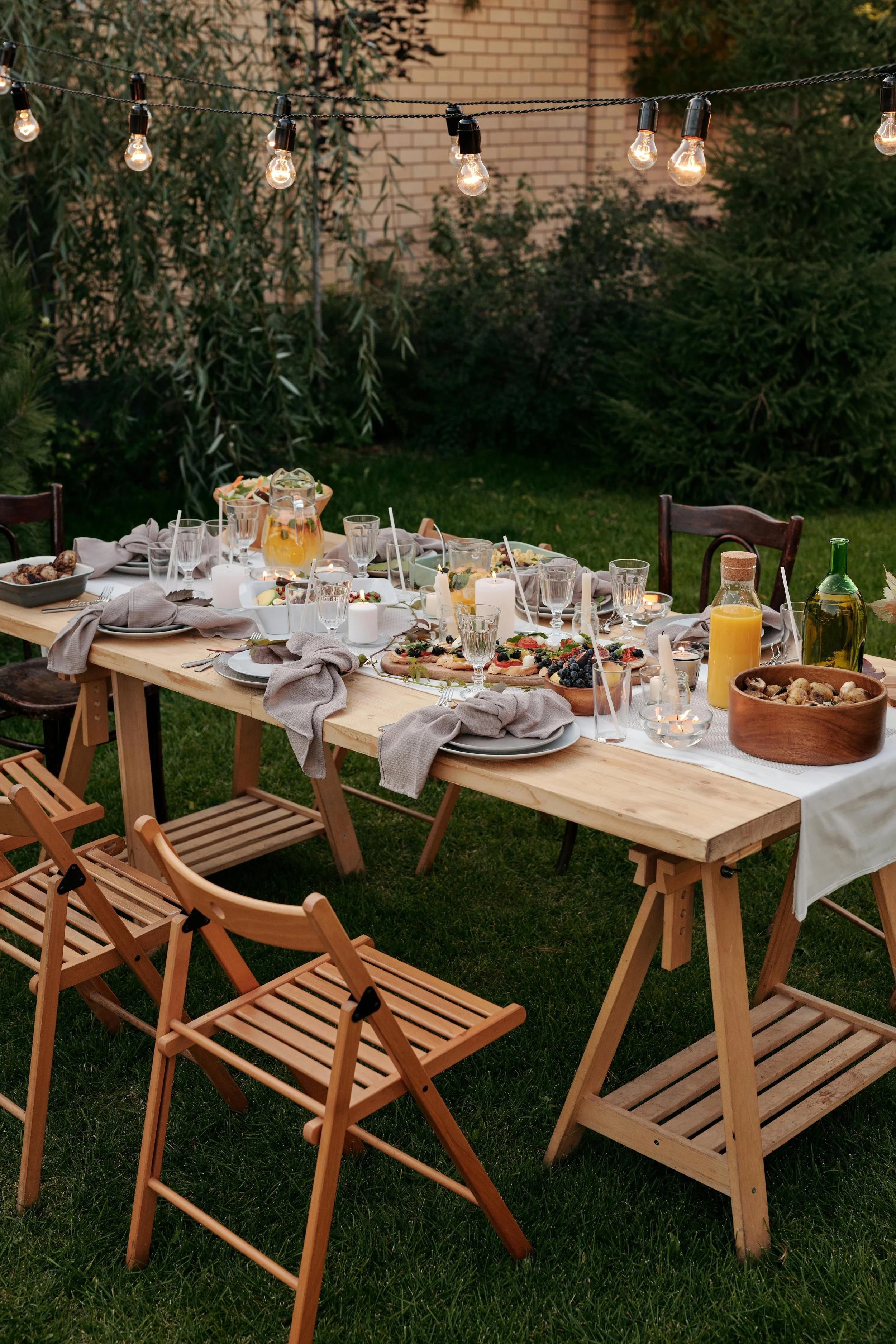 Outdoor dinner table set for a gathering, decorated with food, drinks, and candles, under string lights on a grassy backyard with trees and a brick wall in the background.