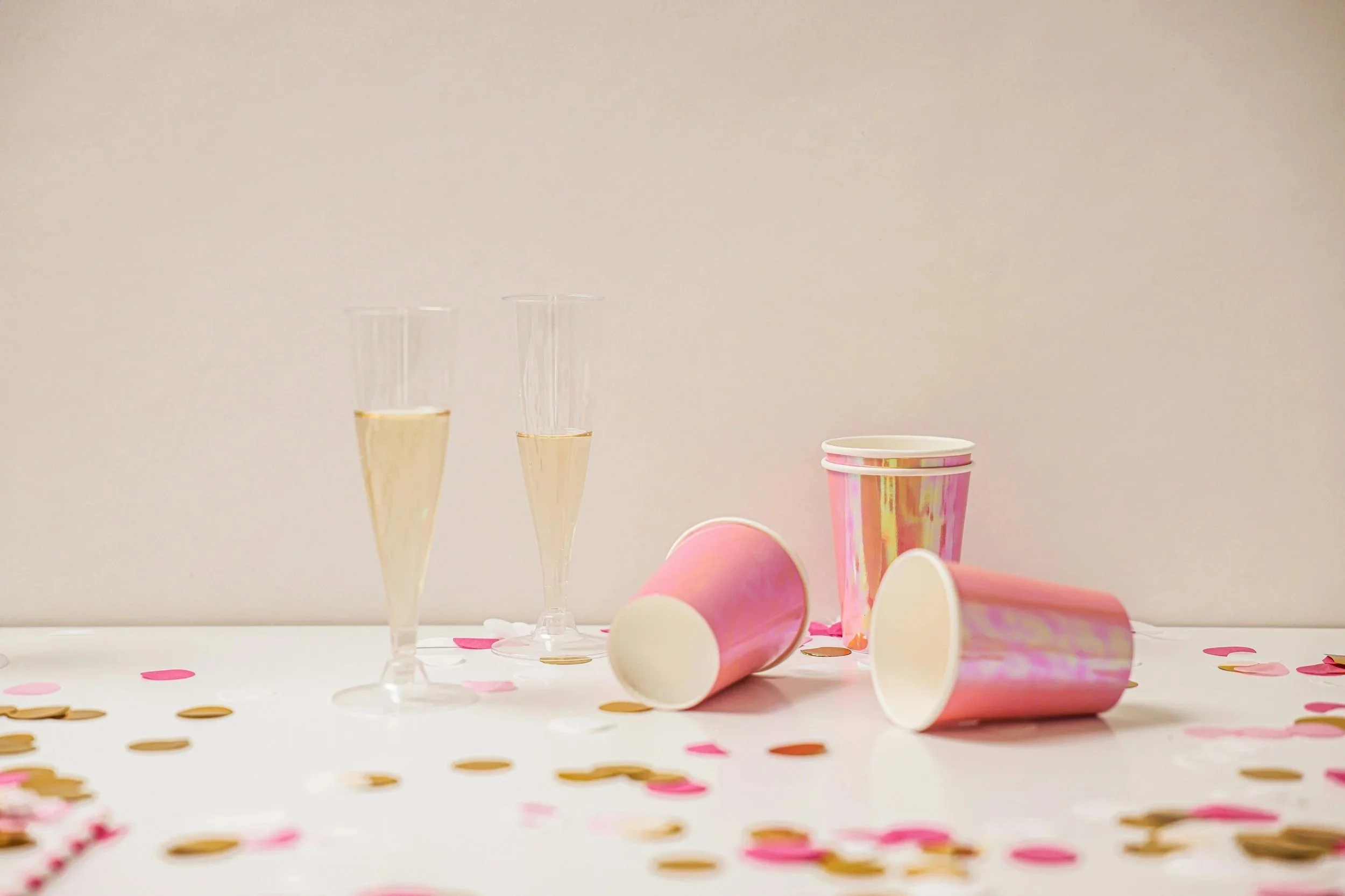 Party scene with pink and holographic cups, some upright and some lying on their sides, two champagne flutes filled with bubbly, and scattered pink and gold confetti on a white surface against a plain background.