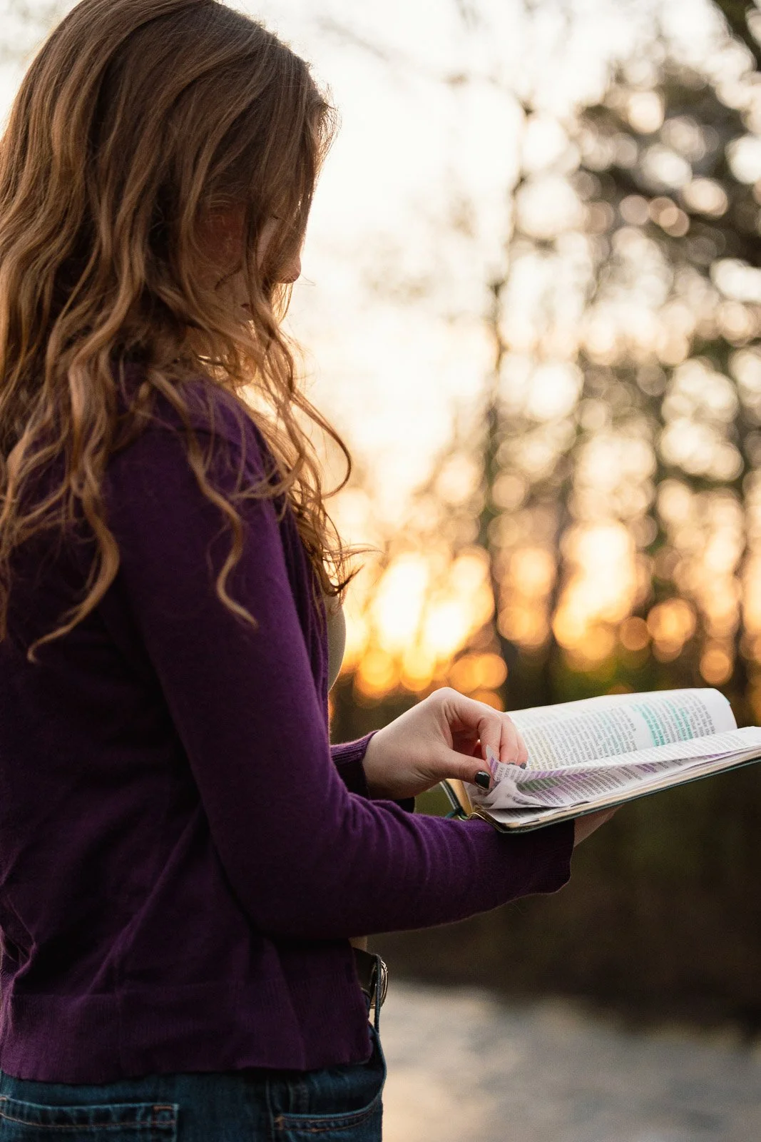 Teenage girl stands in the woods reading a Bible at sunrise.