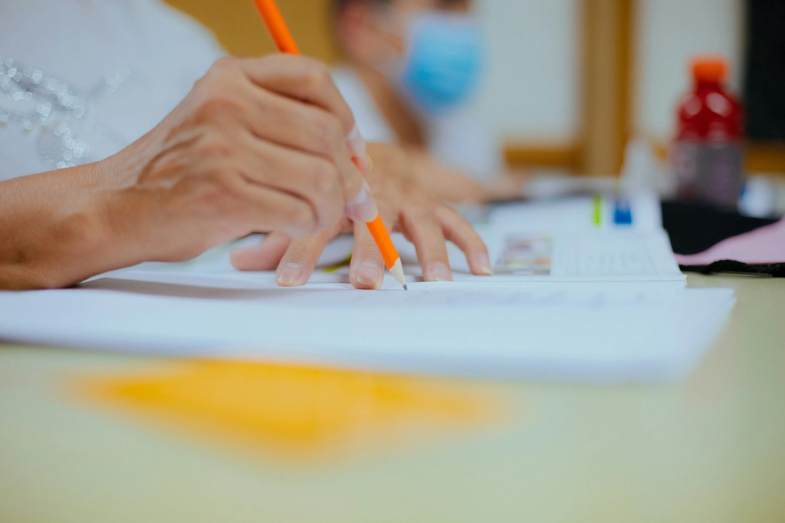 Close-up of a person's hands writing with a pencil on paper with a blurred individual in the background, wearing a face mask.
