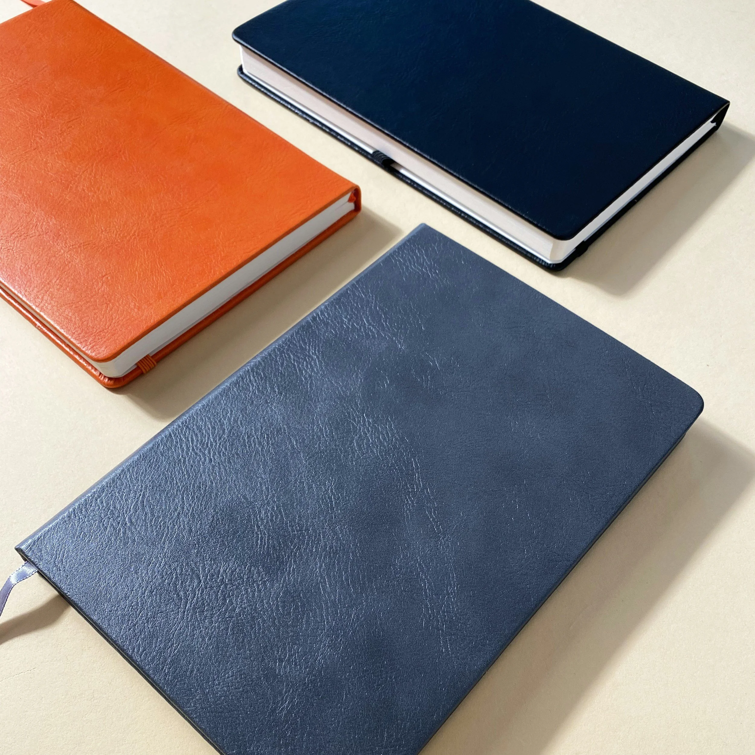 Three closed notebooks or journals with textured covers in orange, navy blue, and dark blue, arranged on a light beige surface.