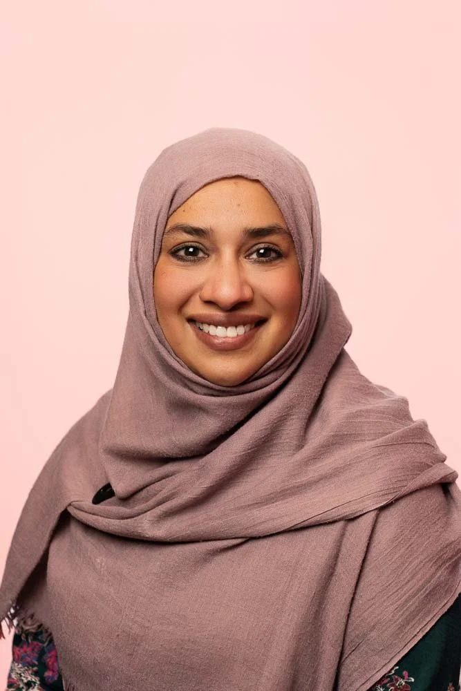 Portrait of a smiling woman wearing a light purple hijab against a pink background.