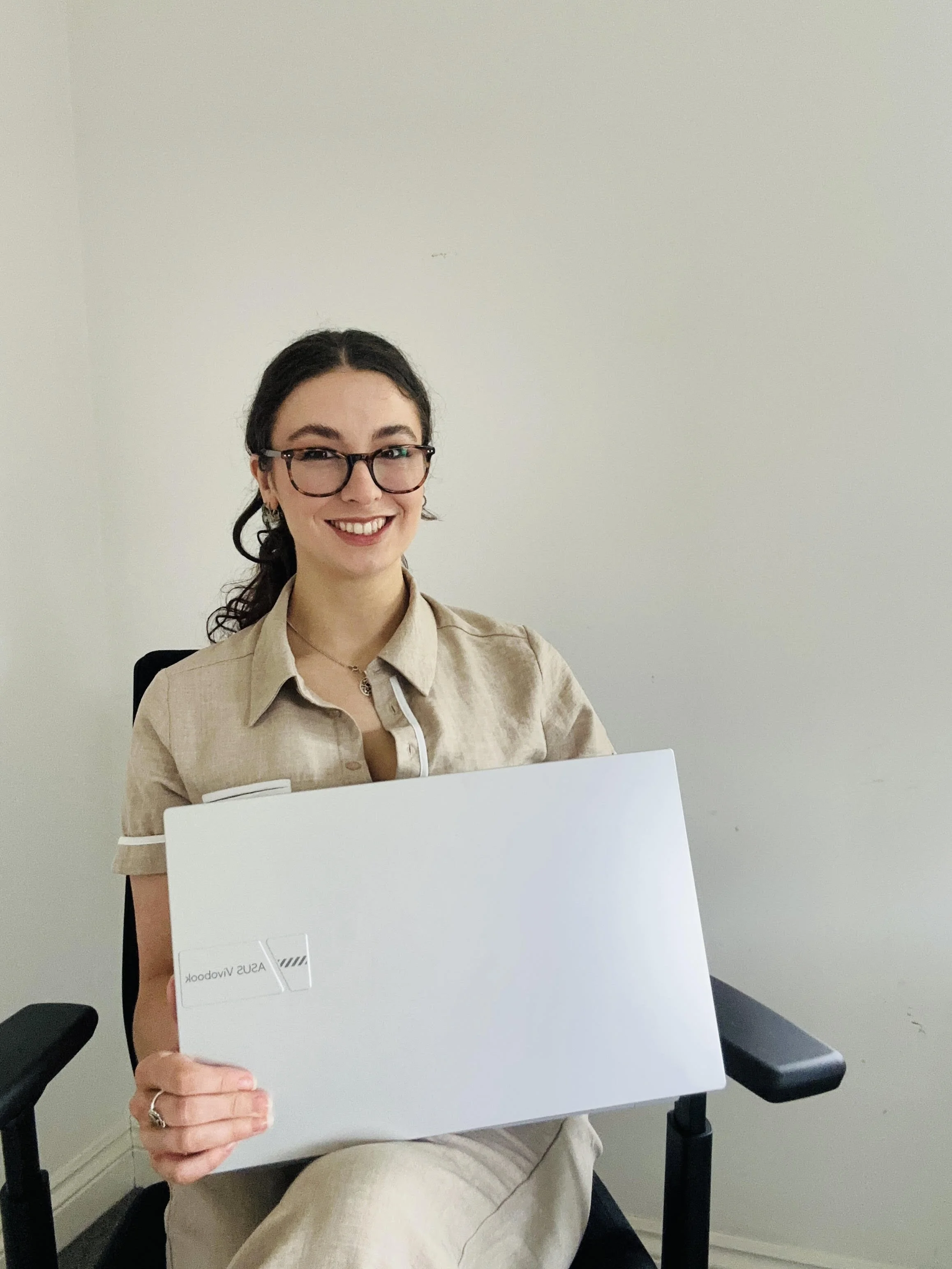 A woman with glasses smiling while sitting in a chair and holding a white laptop or tablet that says 'ASUS VivoBook' on it, in a room with a plain white wall.