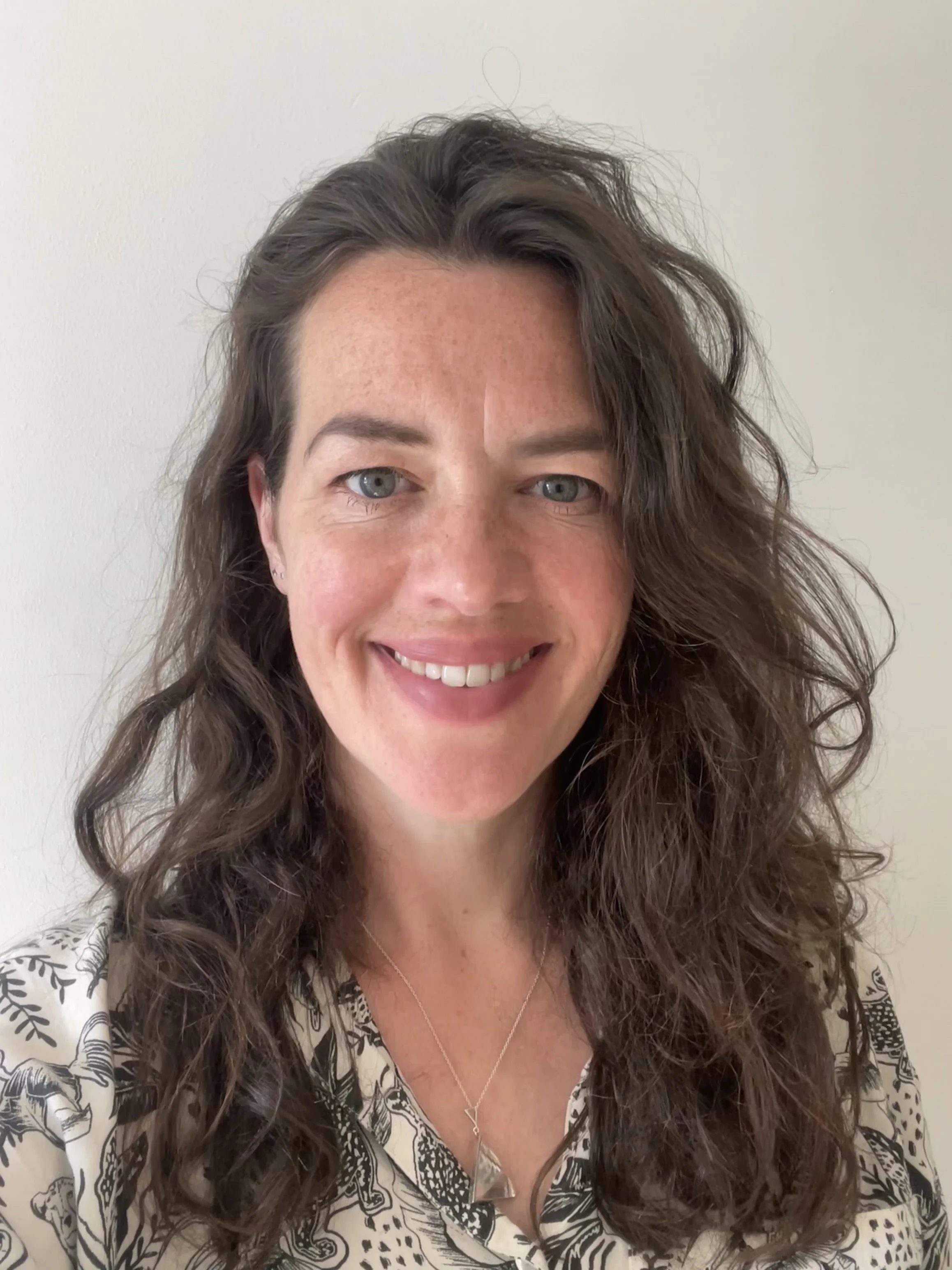 Close-up of a smiling woman with long, curly brown hair and blue eyes, wearing a patterned blouse and a silver necklace, against a plain white wall.