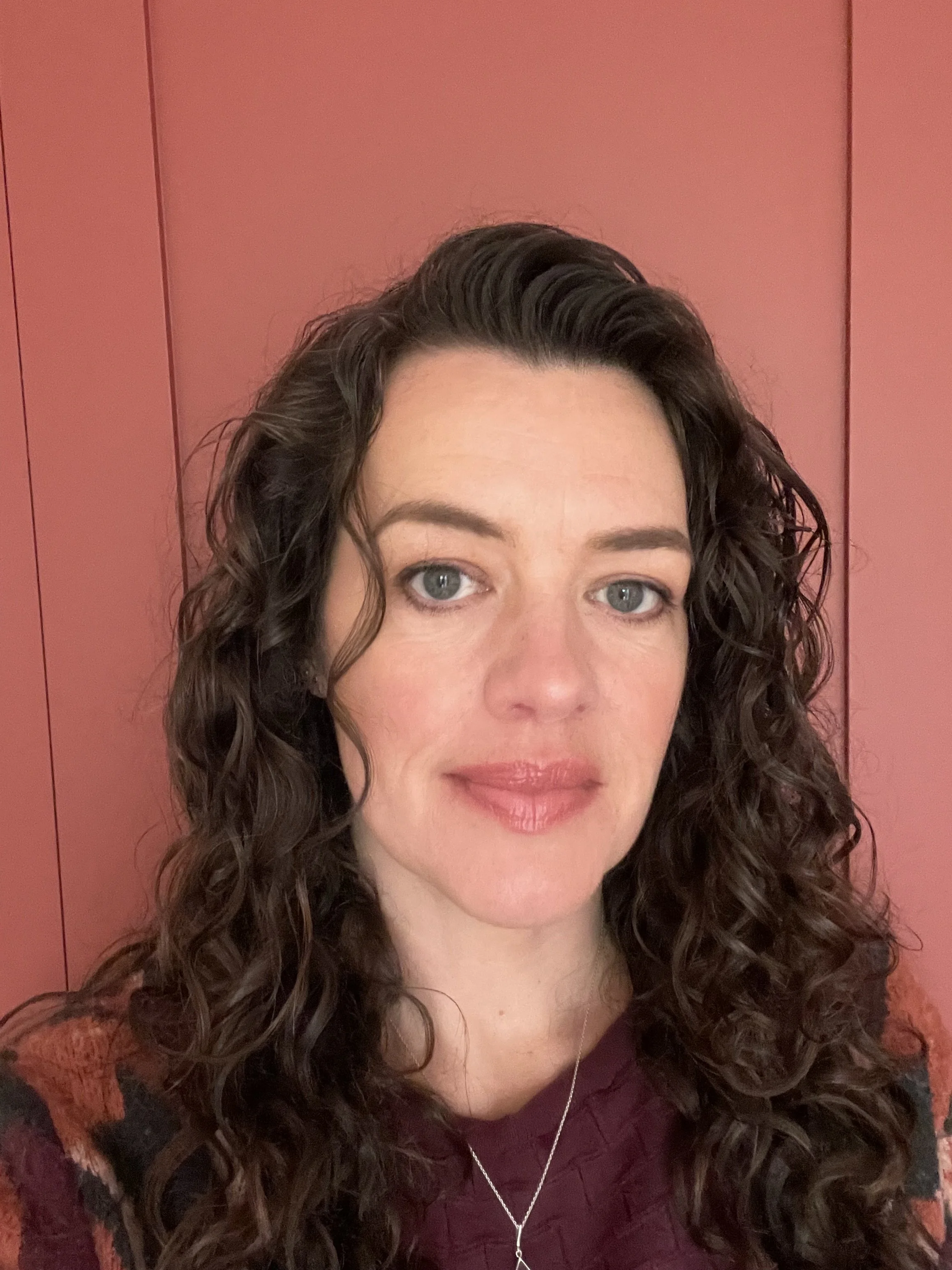A woman with curly brown hair, blue eyes, and wearing a maroon top, standing in front of a pink wall.
