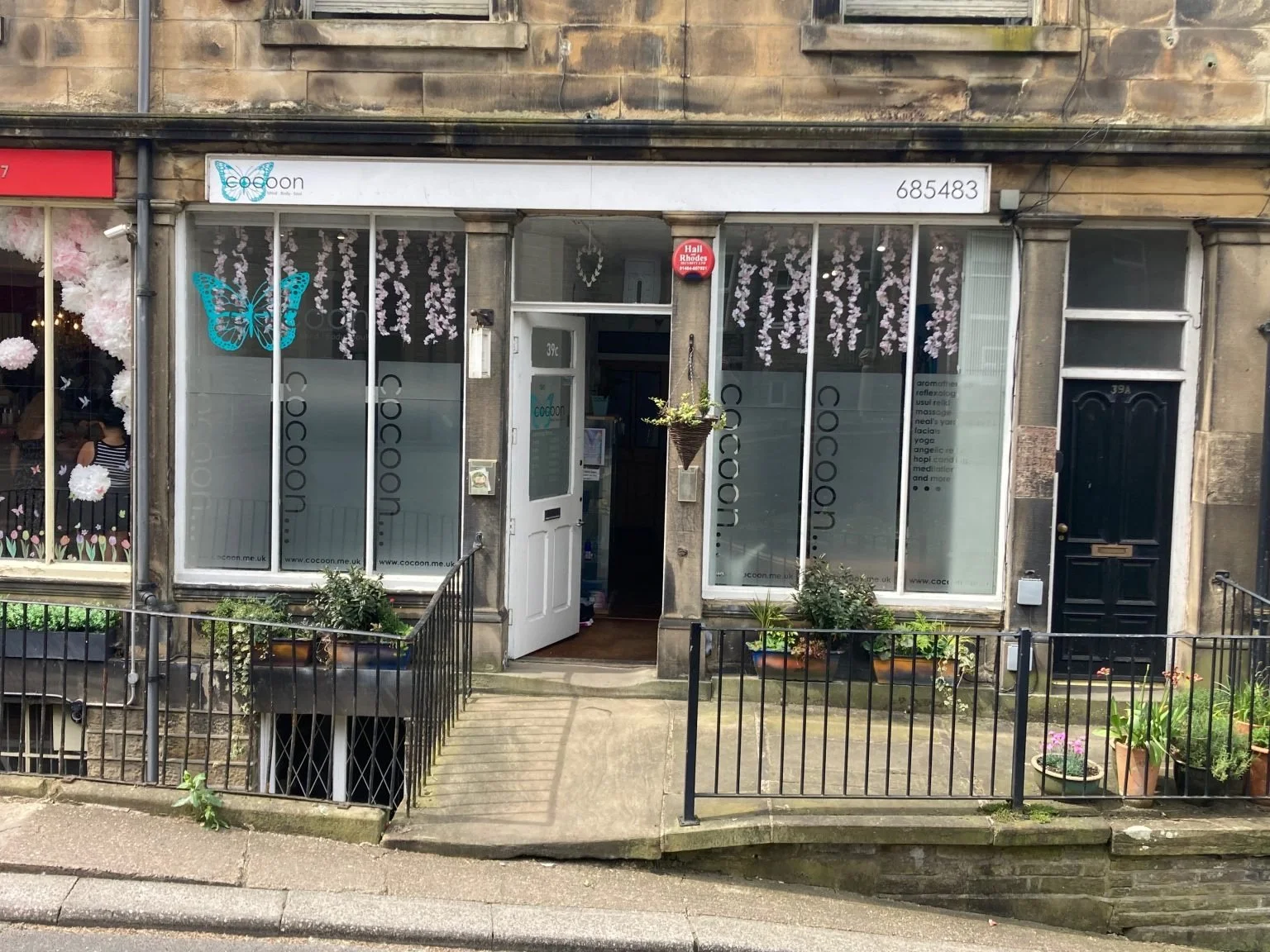 Front view of the storefront for Cocoon, a wellness and massage studio, with pink flower decorations on the large windows. The entrance has a white door with the number 39c, a hanging flower basket, and potted plants on the steps and in front. The signage displays the name Cocoon with a butterfly logo.