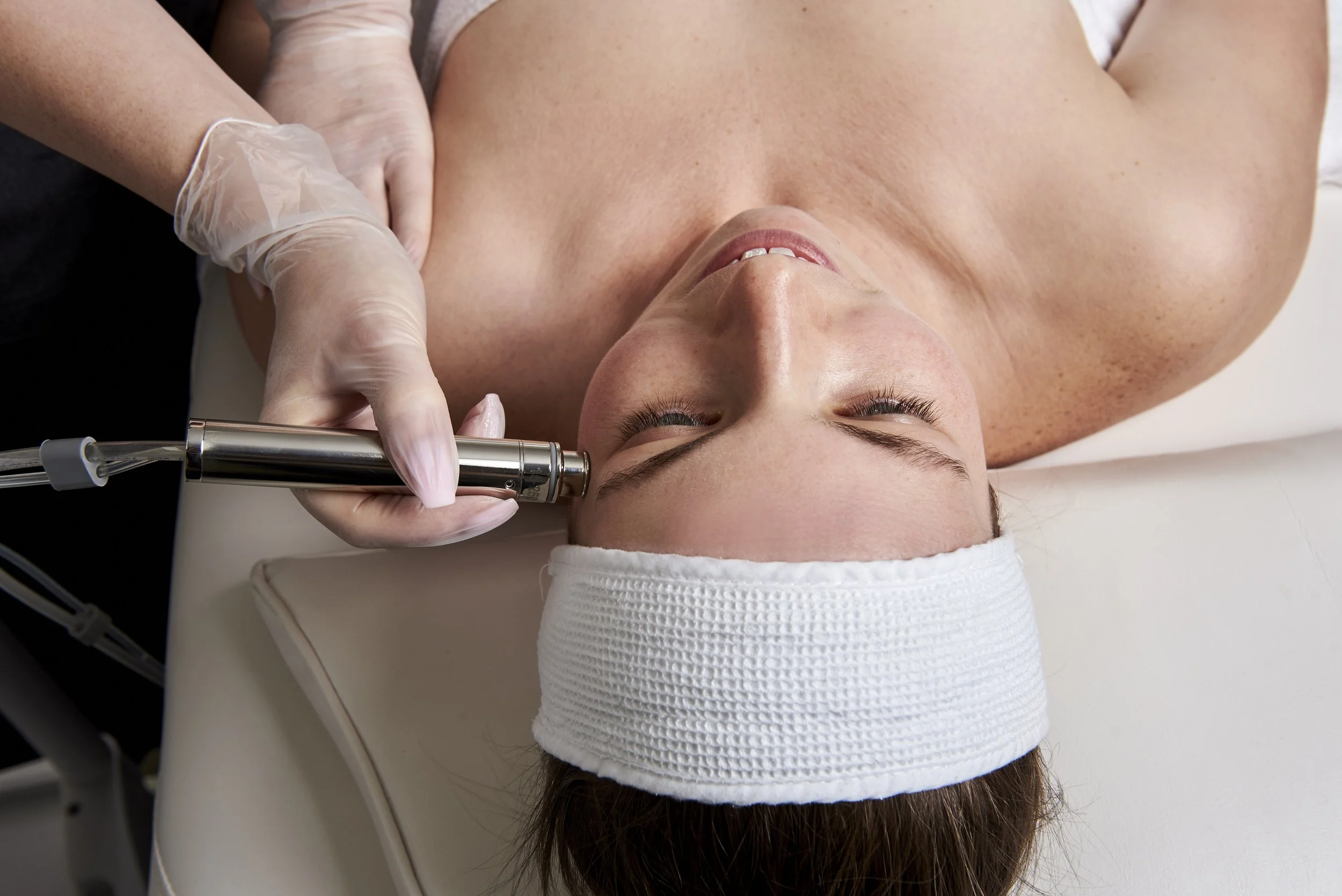 A woman lying on a treatment bed with a white headband, receiving a cosmetic or facial treatment using a handheld device from a professional wearing gloves.
