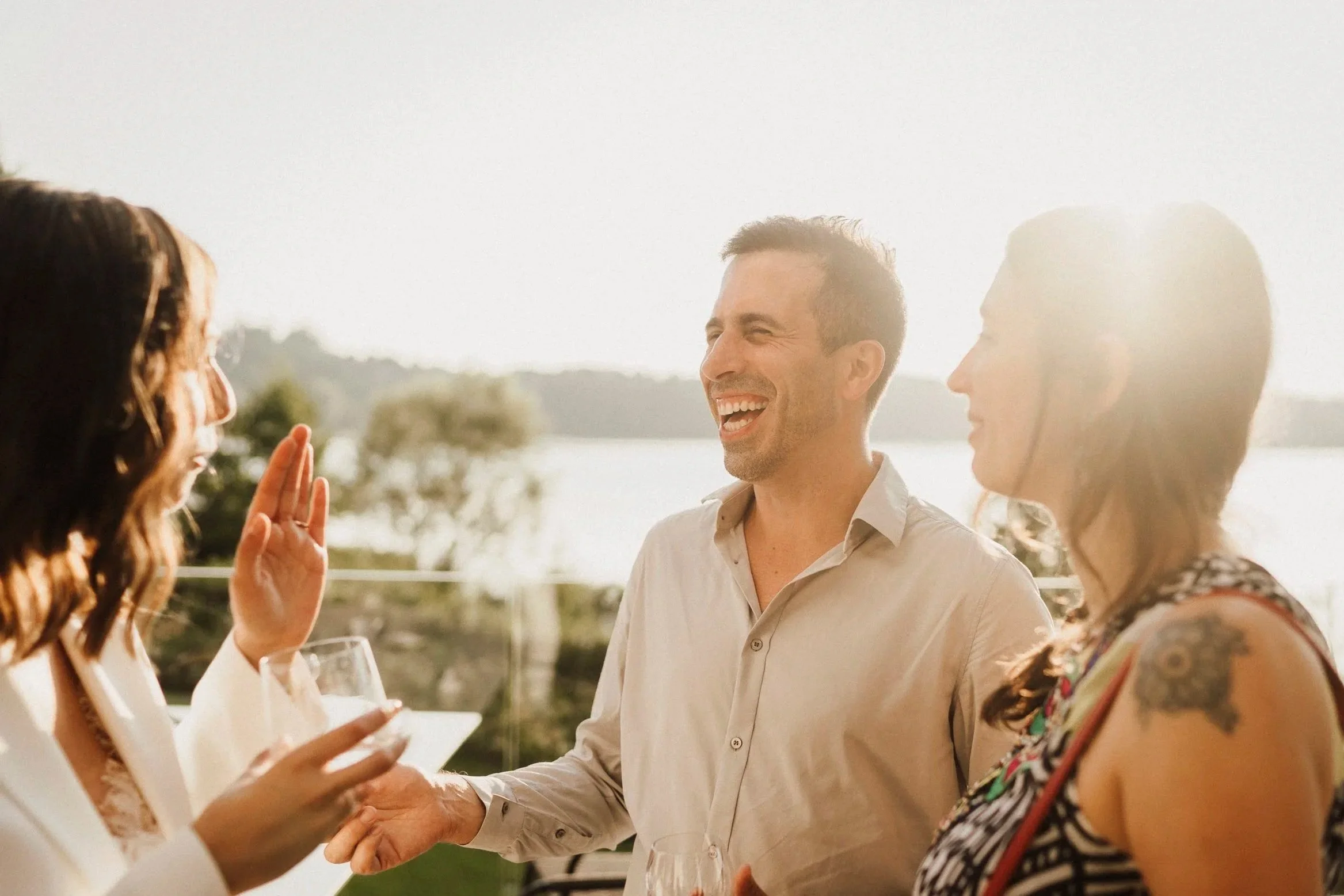 A man holding a glass laughs with a few other people at an outside social gathering