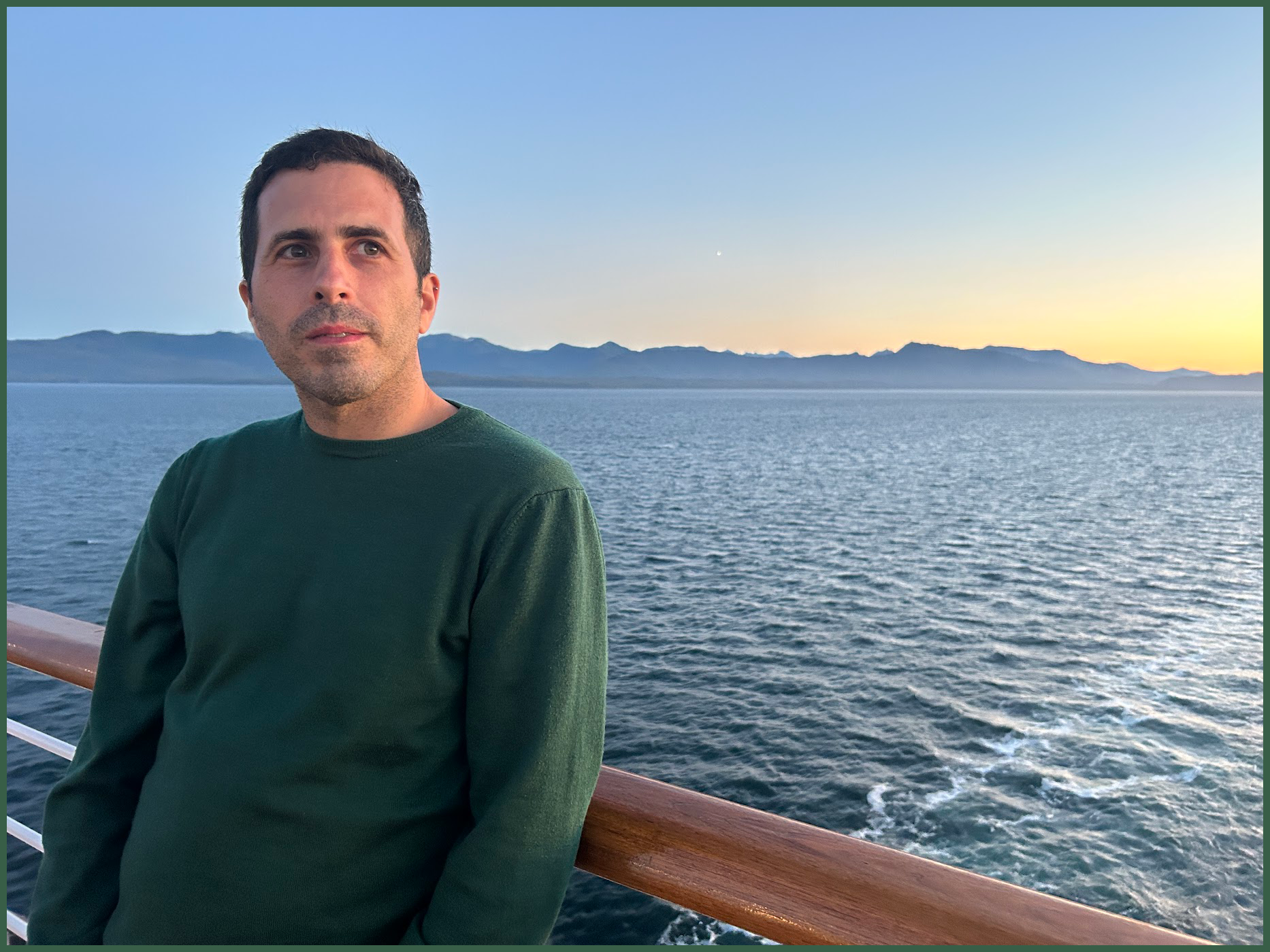 A man in a green sweater standing on a boat deck with a body of water and mountains in the background during sunset.