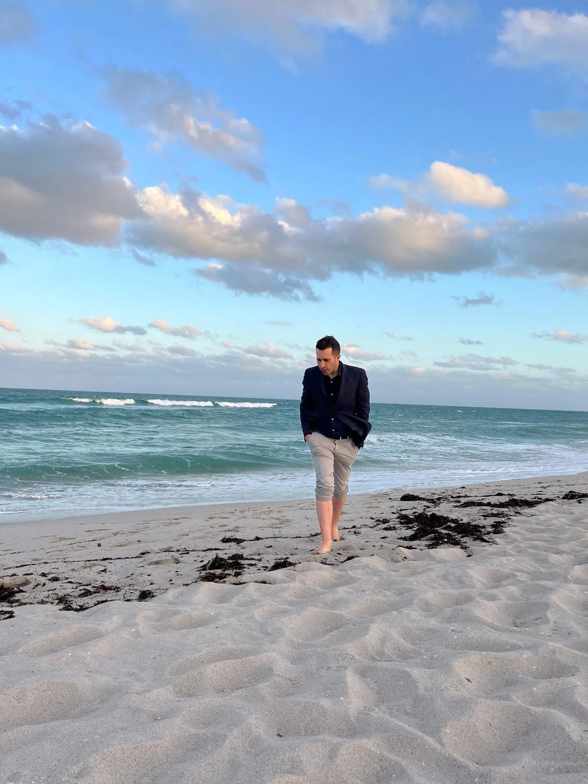 A man wearing rolled up trousers walks on the beach with his hands in his pockets