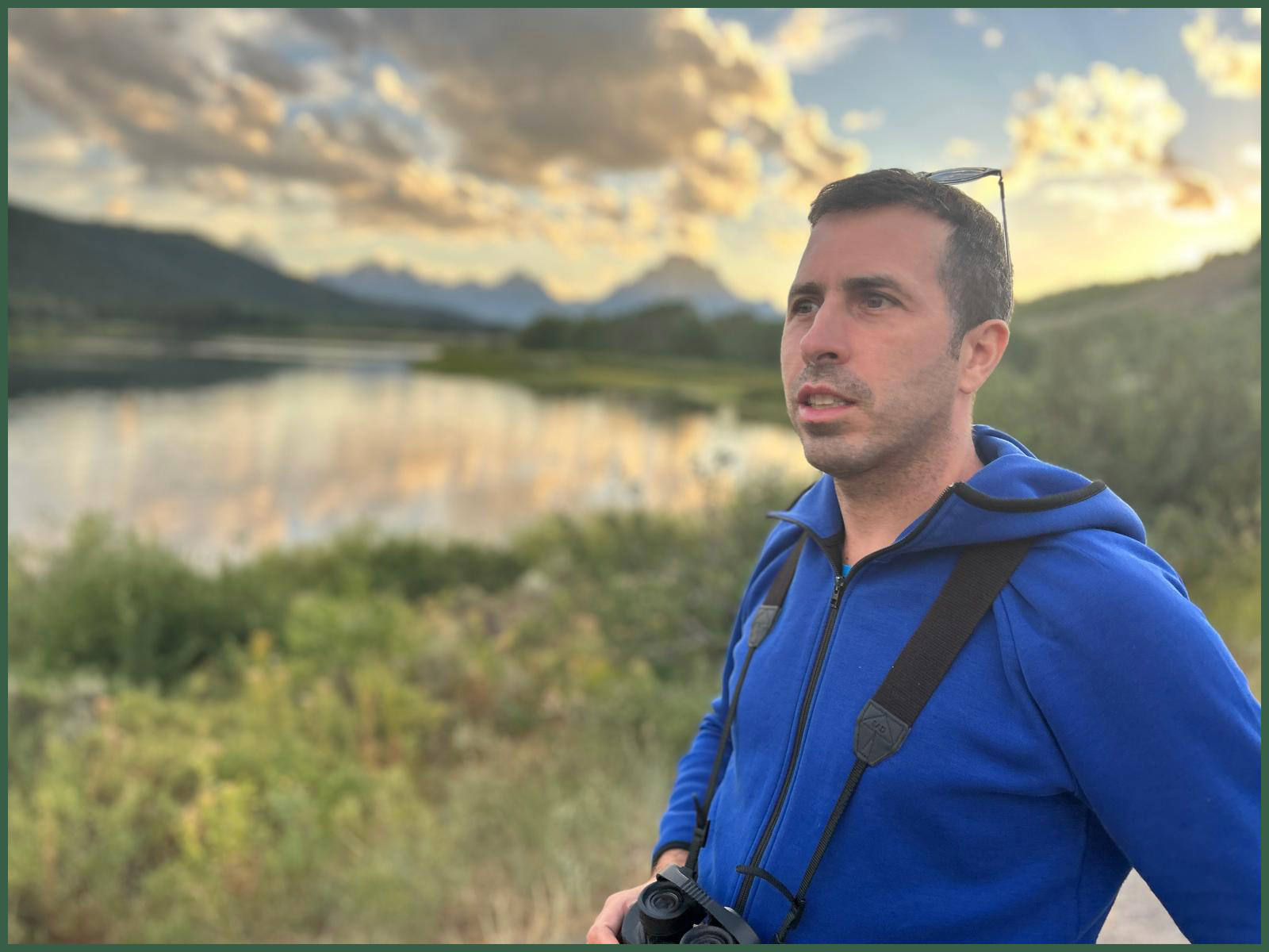 A man in a blue jacket and carrying binoculars stands outdoors near a body of water during sunset, with mountains and clouds in the background.