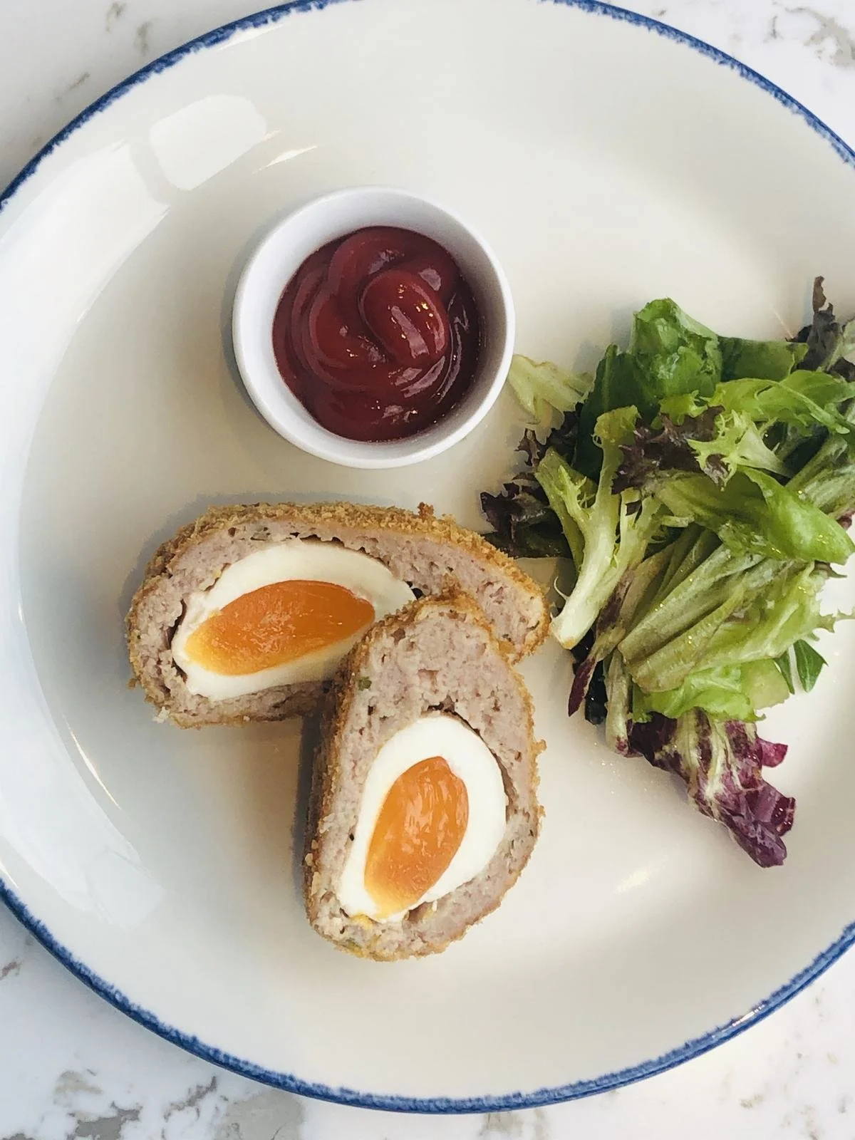 A scotch egg cut in half showing a soft yolk, with salad and ketchup.