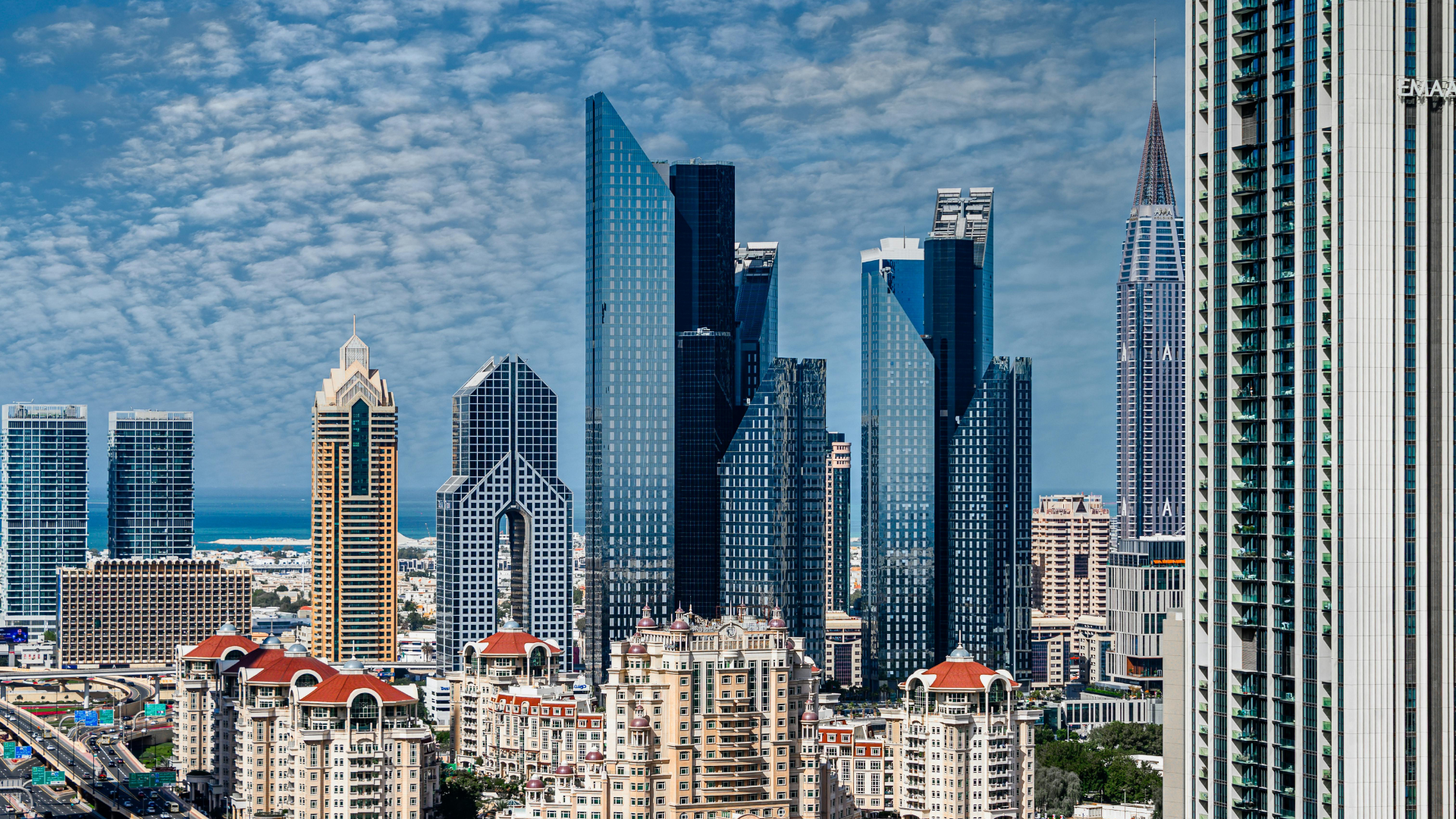 City skyline with tall skyscrapers, some having reflective glass exteriors, and shorter residential buildings with red roofs, under a partly cloudy blue sky.