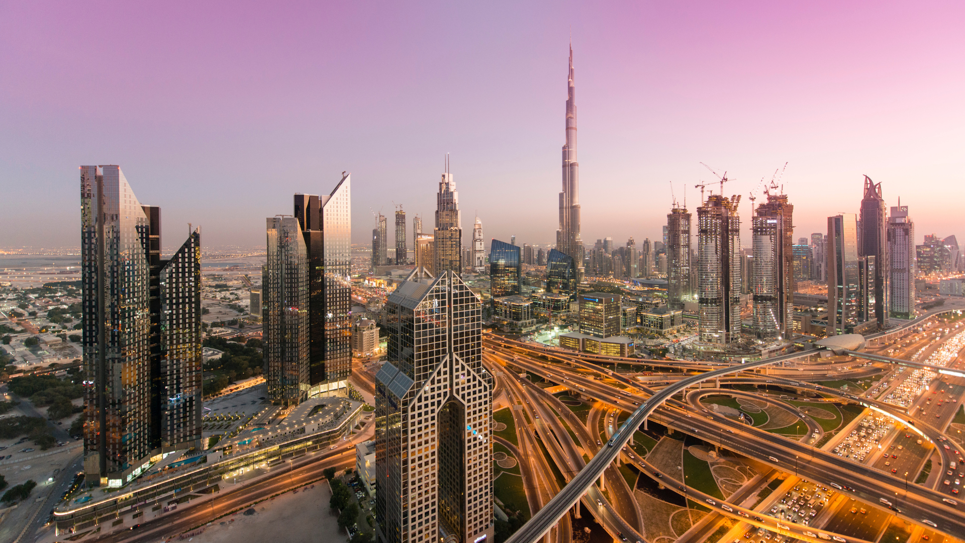Skyline of Dubai featuring the Burj Khalifa at sunset with city lights and construction cranes.