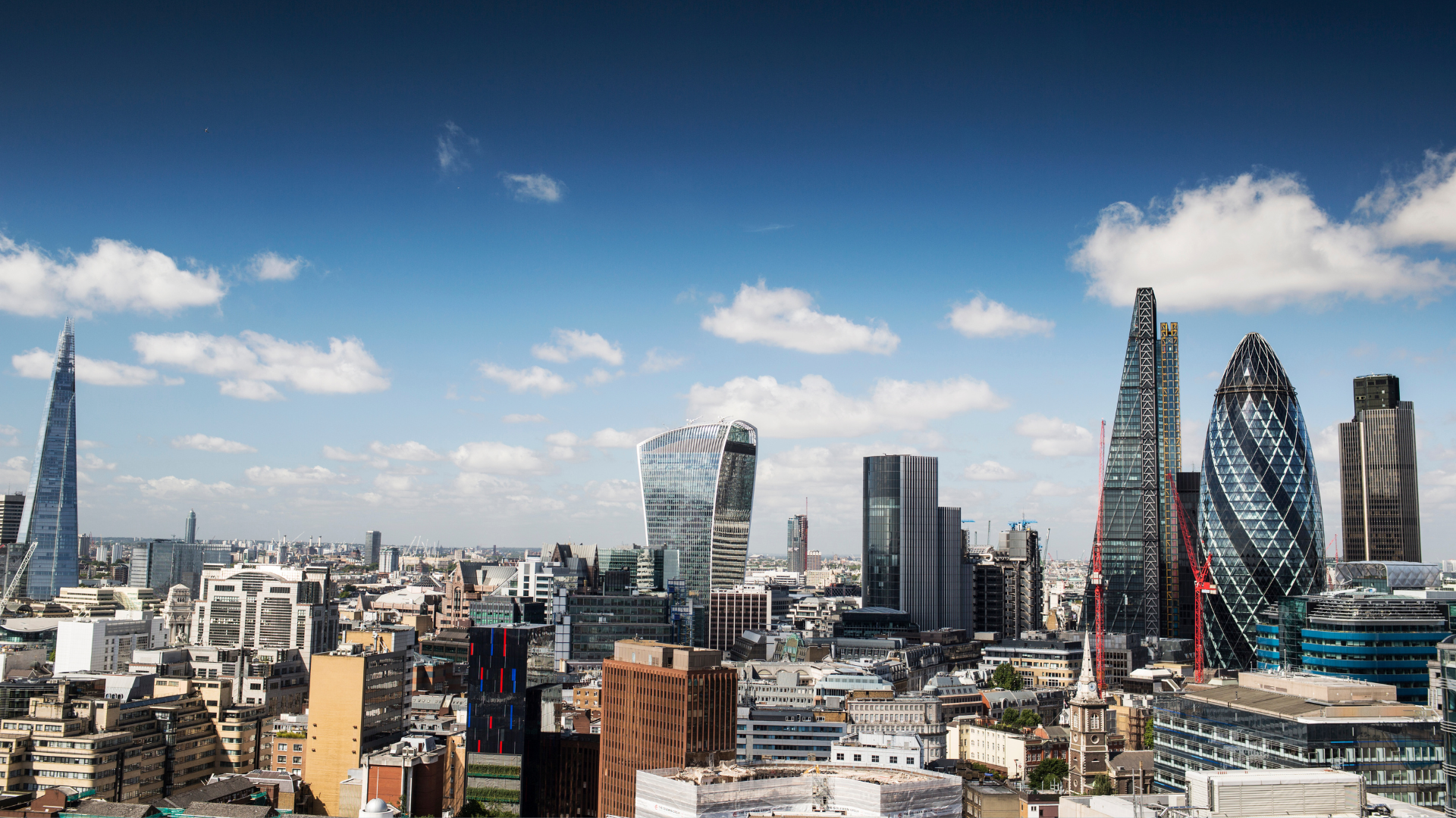 Panoramic view of London's skyline on a clear day with modern skyscrapers, including The Shard and the buildings in the City of London, under a blue sky with scattered clouds.