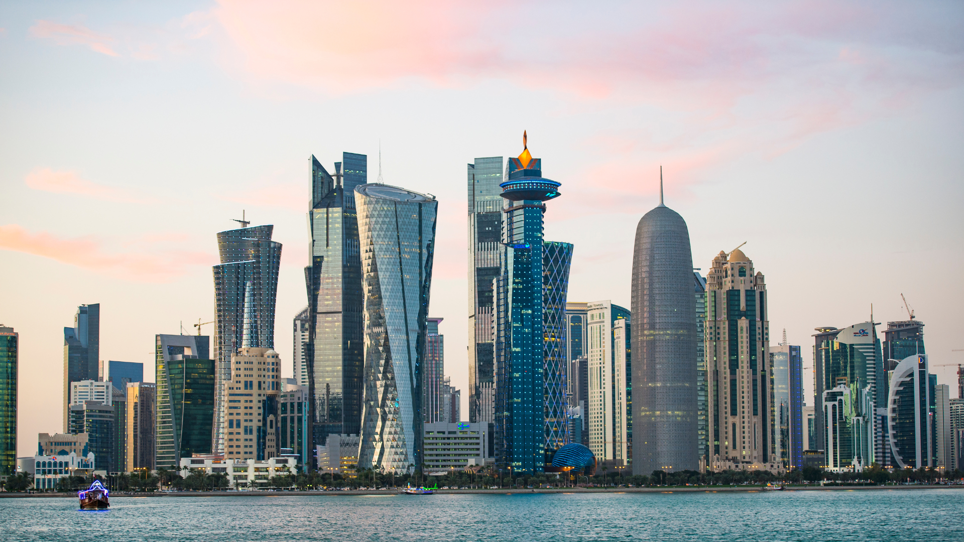 City skyline at dusk with modern skyscrapers, including the twisting Cayan Tower and the cylindrical Emirates Towers, viewed across a body of water with boats, under a pink and blue sky.