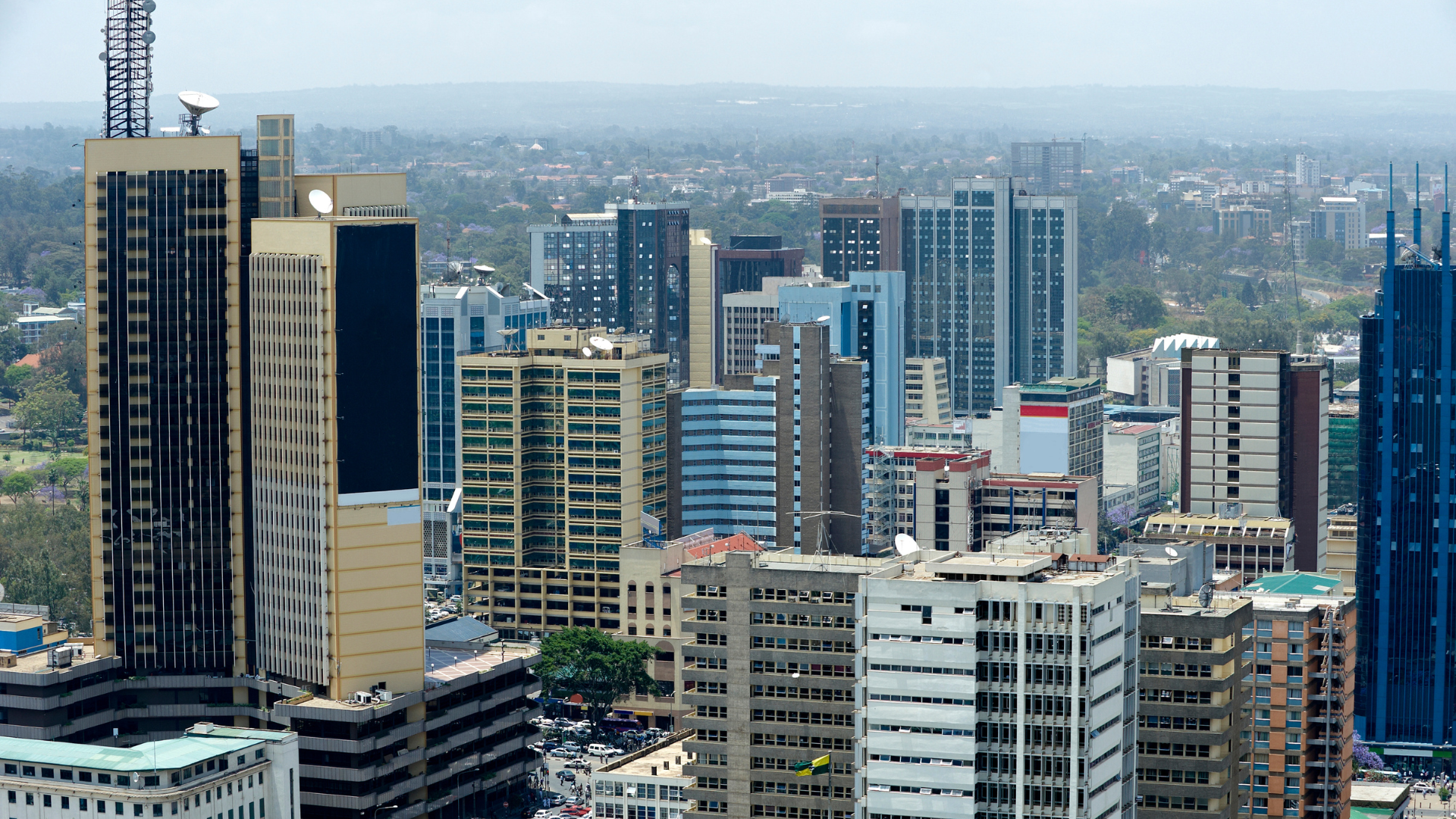 Cityscape with numerous high-rise office buildings and skyscrapers, some with satellite dishes and antennas on rooftops, background of distant hills and a cloudy sky.