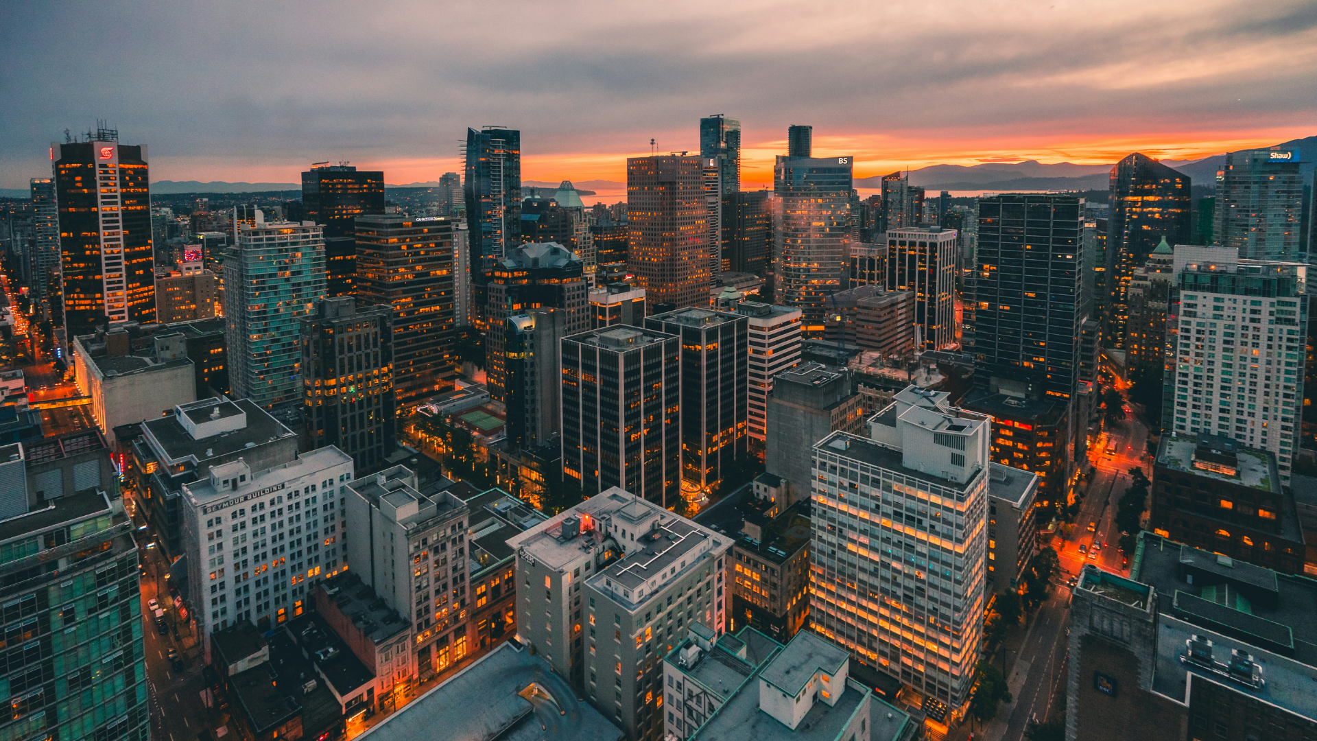 A city skyline at sunset with illuminated skyscrapers and buildings across the urban landscape, with mountains in the background and a colorful sky.