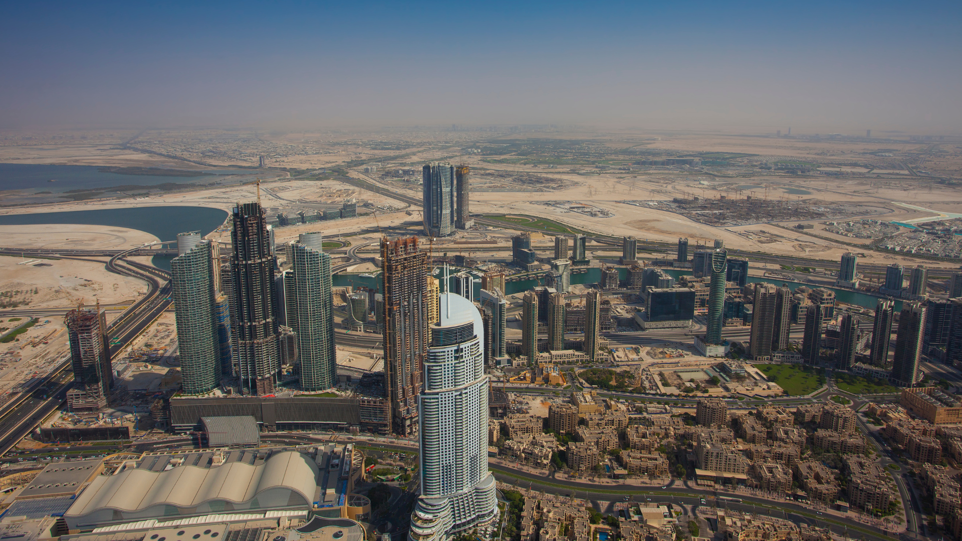 Aerial view of Dubai skyline featuring skyscrapers, waterways, roads, and construction sites in a desert landscape.