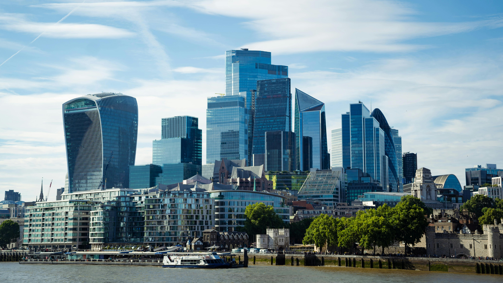 Skyline of London with modern skyscrapers near the River Thames, featuring the Walkie Talkie building and other tall glass buildings under a blue sky with clouds.
