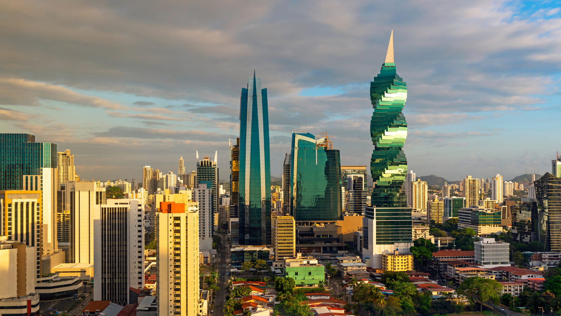 Downtown city skyline with modern skyscrapers, including the twisted green-tinted Templo Mayor building and the sharp, blue-tinted Torre Reforma tower, under a partly cloudy sky.
