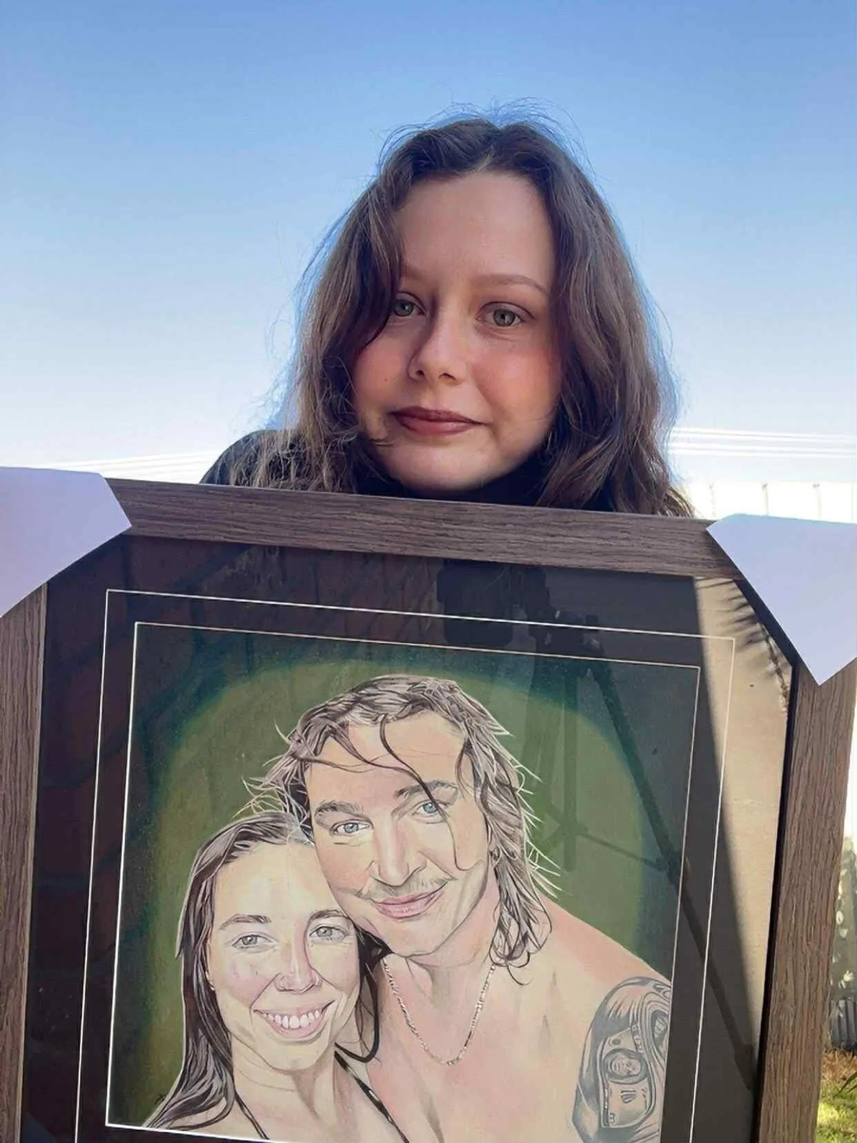 A woman with brown curly hair stands outdoors under a clear blue sky, holding a framed caricature portrait of herself and a man. The caricature depicts both with exaggerated happy expressions, and the background of the portrait is green.