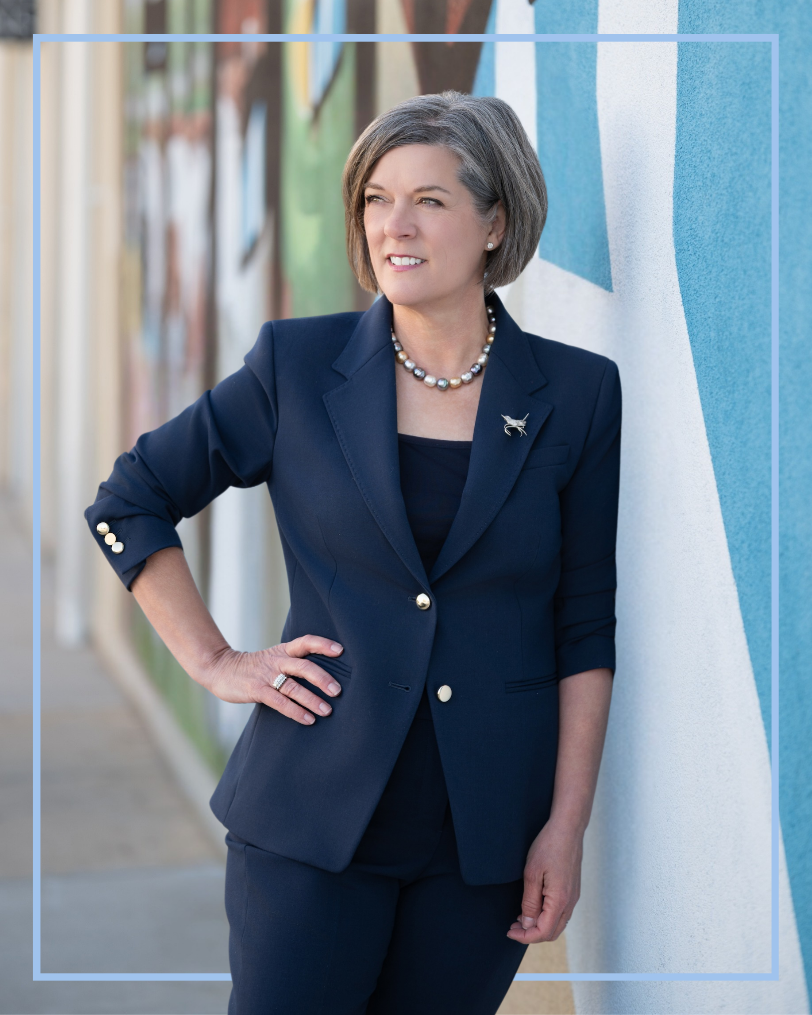 Caroline Avinger, candidate for SC House District 24, leans on a wall with a mural in downtown Greenville. She is wearing a navy blue suit and has her right hand on her hip.