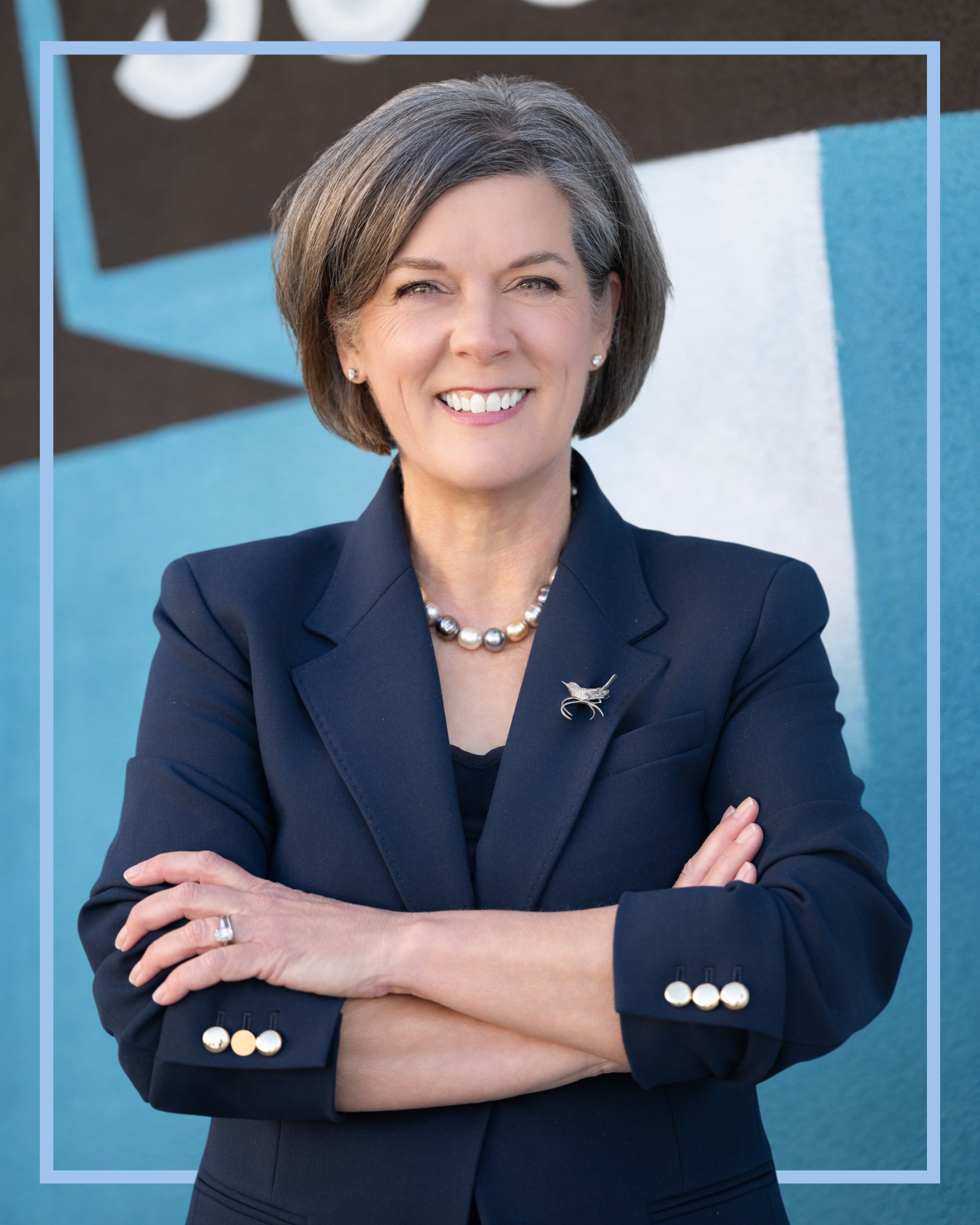 Caroline Avinger, candidate for SC House District 24, leans on a wall smiling with a mural in downtown Greenville. She is wearing a navy blue suit with a Carolina Wren pendant, and has her arms crossed.