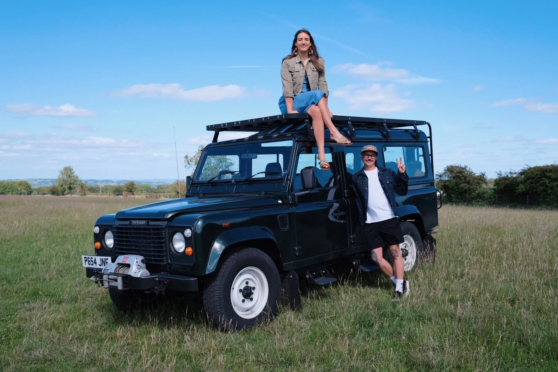 A woman sitting on top of a black Land Rover Defender with two men standing beside it in a grassy field with trees in the background.