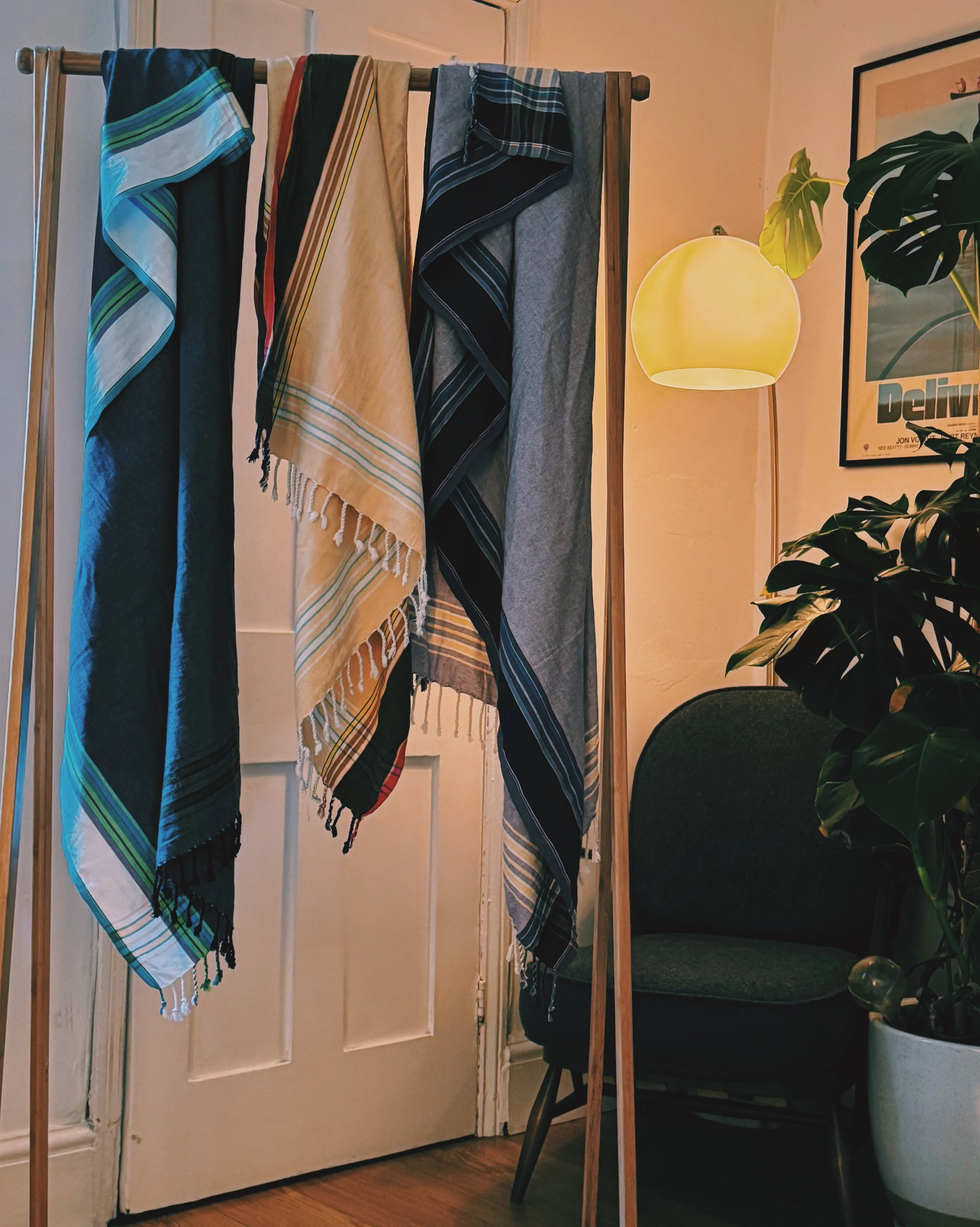 Four towels with different colorful and striped patterns hanging on a wooden stand indoors, with a green chair, a potted plant, a framed poster, and an illuminated lamp in the background.
