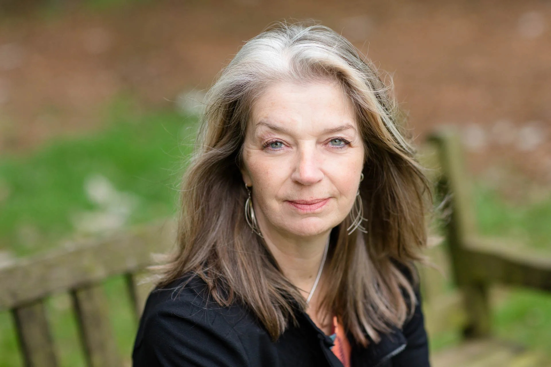 Em Whitfield Brooks a middle-aged woman with long, wavy brown hair and light eye color, smiling softly outdoors with a blurred natural background.