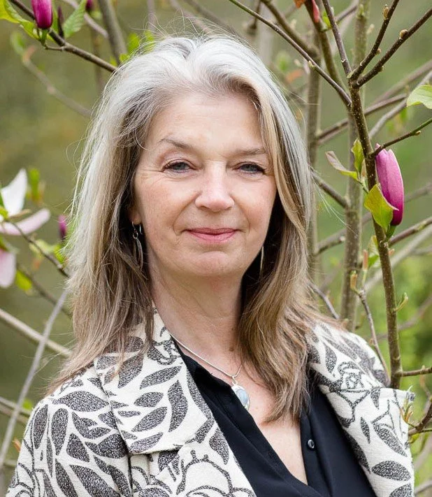 Em Whitfield Brooks, a middle-aged woman with long gray hair, wearing earrings, a necklace, a black top, and a patterned jacket, standing outdoors in front of a blooming tree with pink buds.