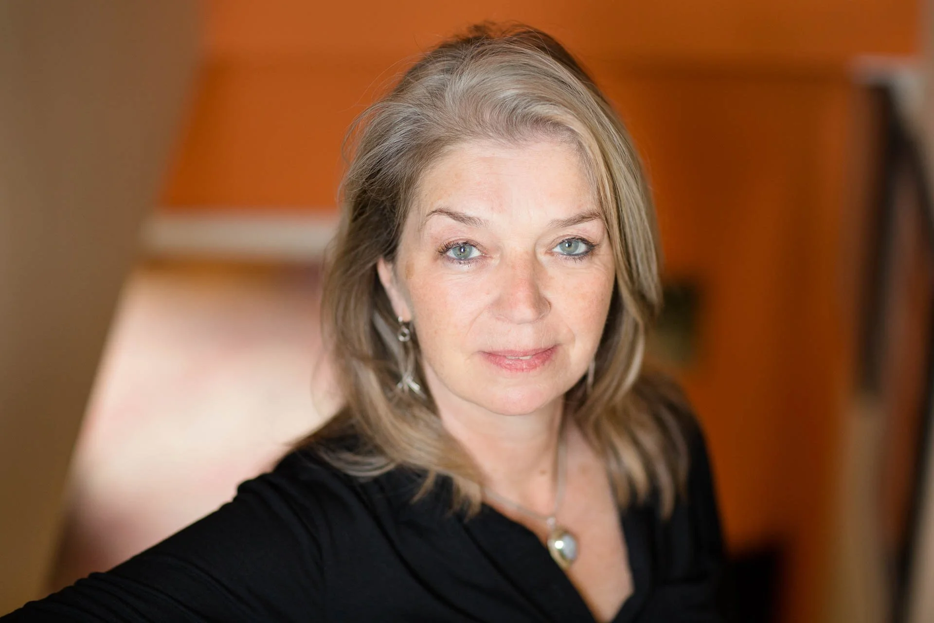 Em Whitfield Brooks, a middle-aged woman with long blonde hair and light blue eyes, wearing a black top, earrings, and a necklace, sitting indoors with a blurred orange background.