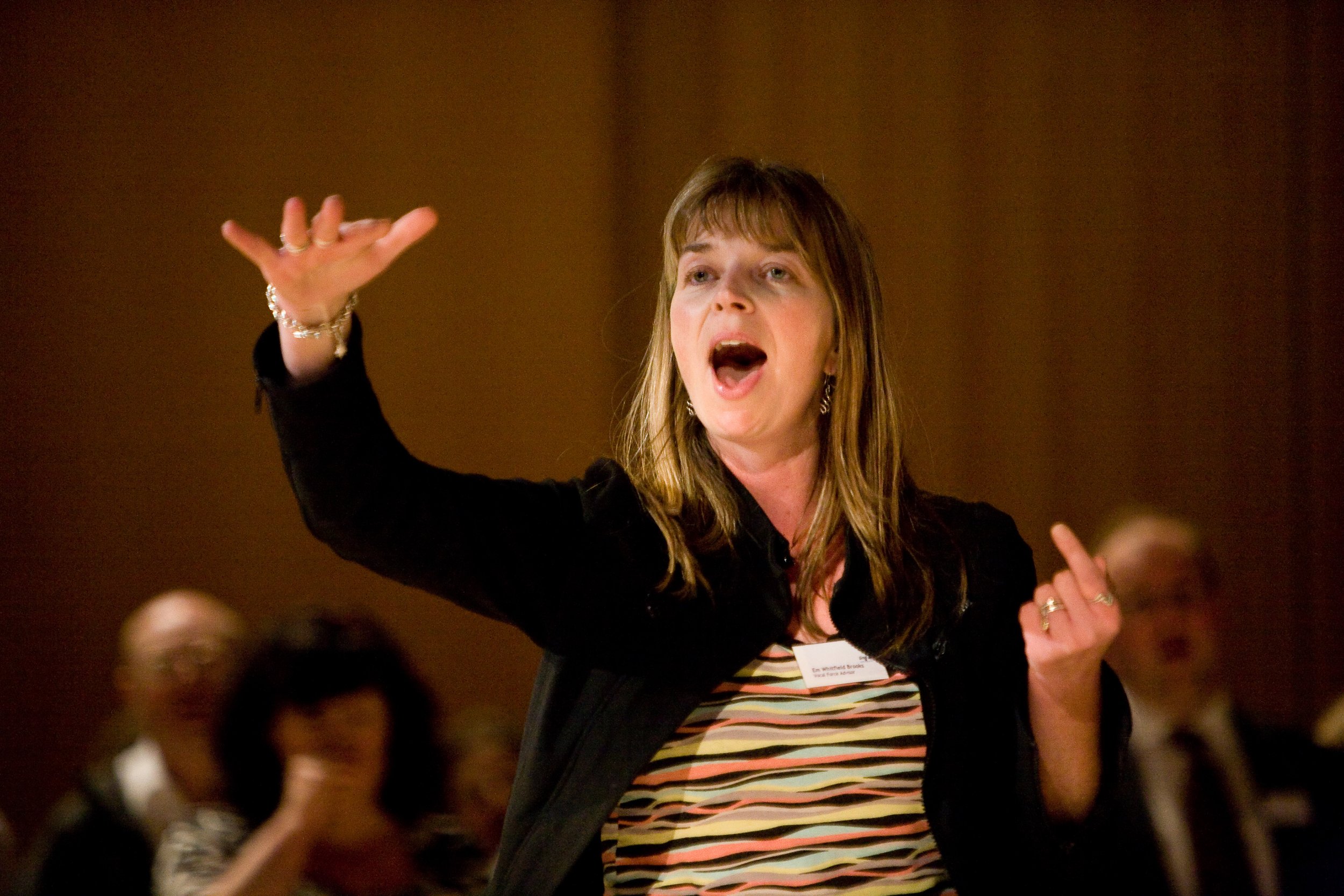 Em Whitfield Brooks, a woman with shoulder-length brown hair gesturing with her right hand while speaking, wearing a colourful striped top and black jacket, with a neutral brown background and singing workshop members in the background.
