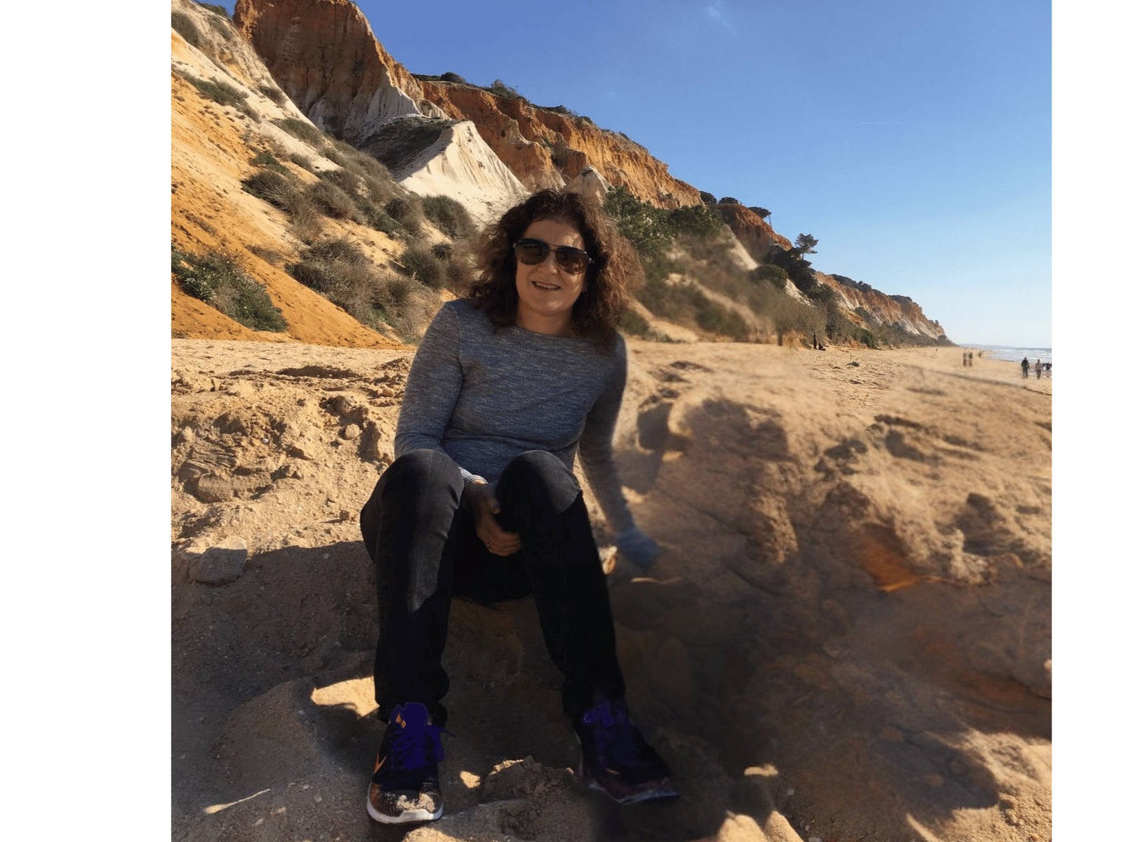 A woman sitting on a sandy beach with colorful cliffs in the background, wearing sunglasses, a gray long-sleeve shirt, black pants, and purple shoes, smiling at the camera.