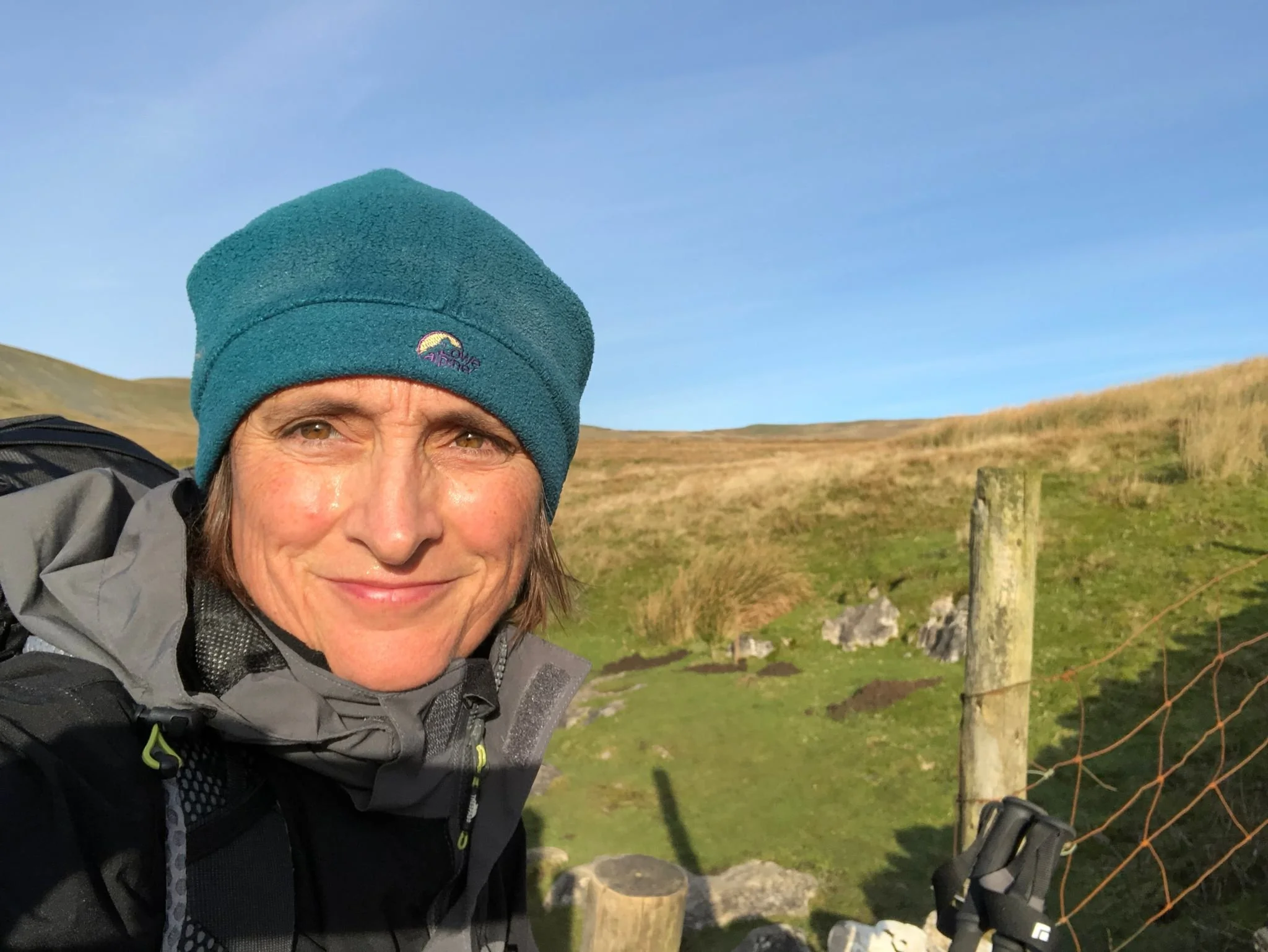 Close-up of a woman's face outdoors, wearing a teal beanie with a logo, black jacket, and carrying a backpack, with grassy hills and wooden fence in the background.