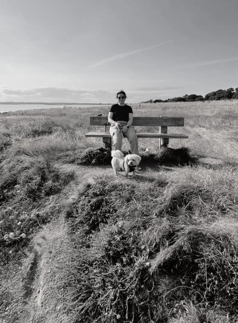 photo of woman sitting on a bench with a small white dog on a lead with sea in background on a clifftop