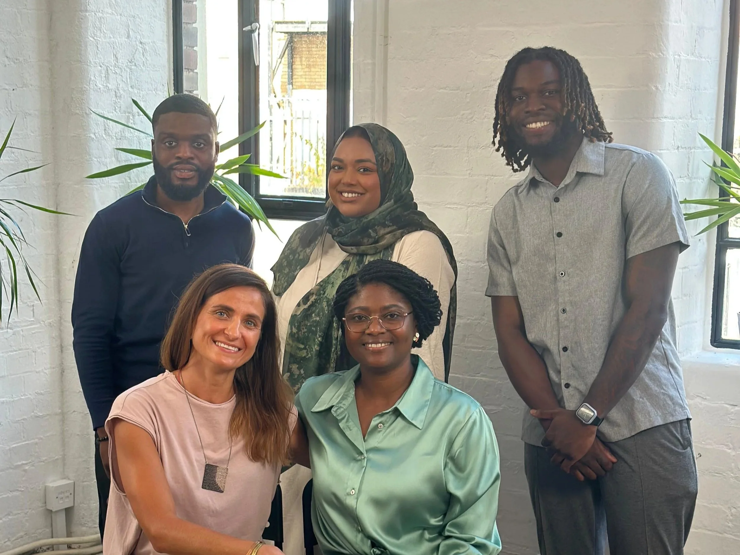 A group of six diverse people posing together indoors near windows and green plants, smiling at the camera.