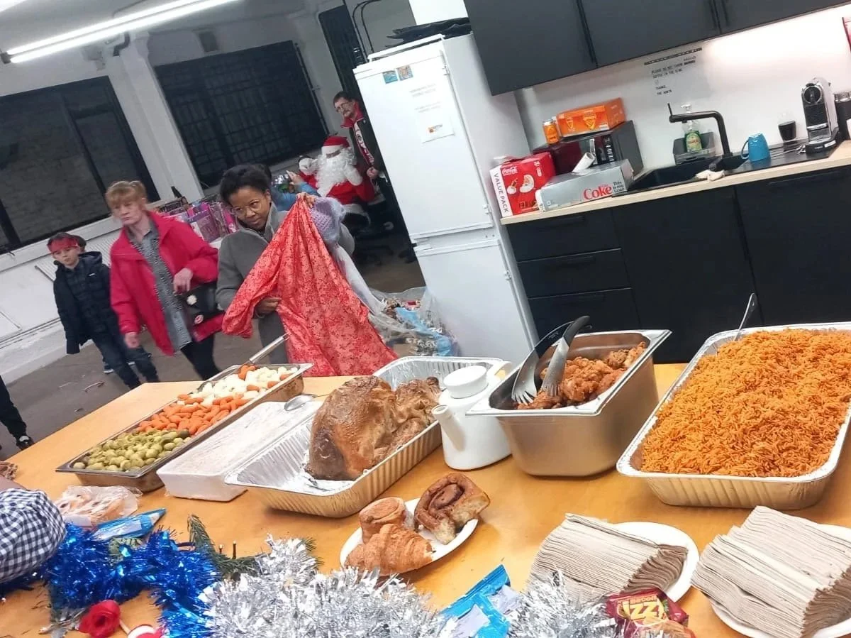 A holiday party buffet table with various dishes including rice, chicken, vegetables, bread, and drinks. People are in the background, with some dressed in Christmas attire, including a person in a Santa costume and others in sweaters, in a room decorated for the holidays.