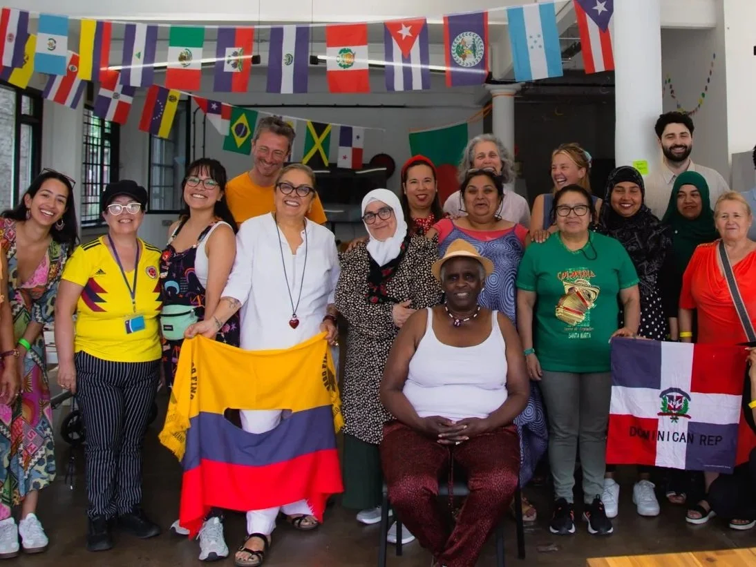 Group of diverse people smiling, celebrating, with international flags hanging from ceiling, some holding flags, in a multicultural indoor setting.