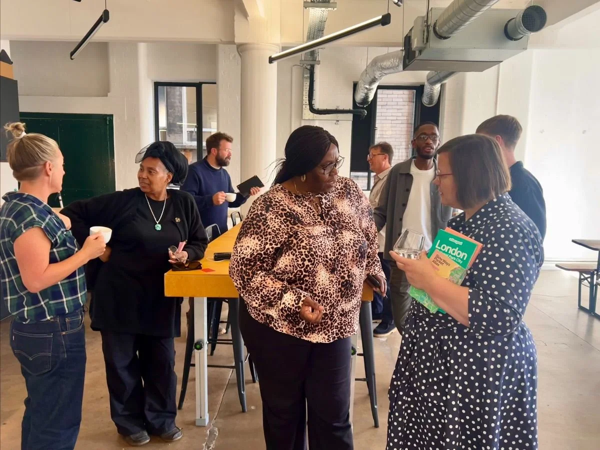 A diverse group of people are gathered indoors, engaging in conversation during what appears to be a social or networking event. Some hold cups and pamphlets or notebooks.